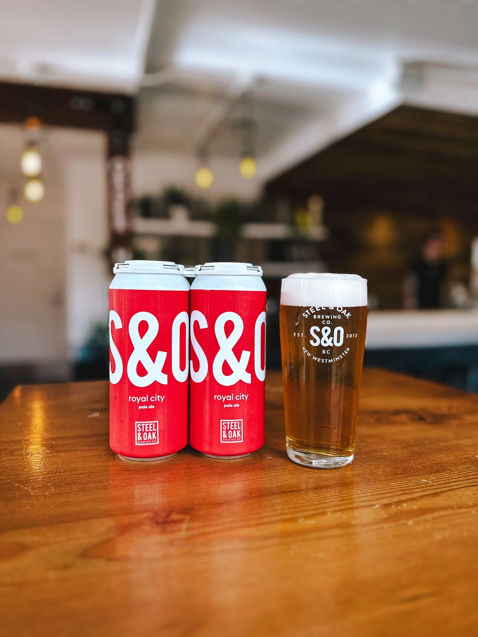 Two cans of Steel & Oak Royal City pale ale beer and a pint glass filled with beer on a wooden table in a bar.