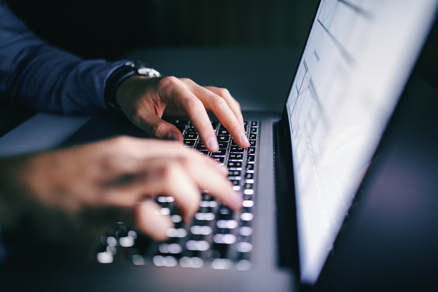 Close-up of a person typing on a laptop keyboard with illuminated keys, seen in a dimly lit environment.