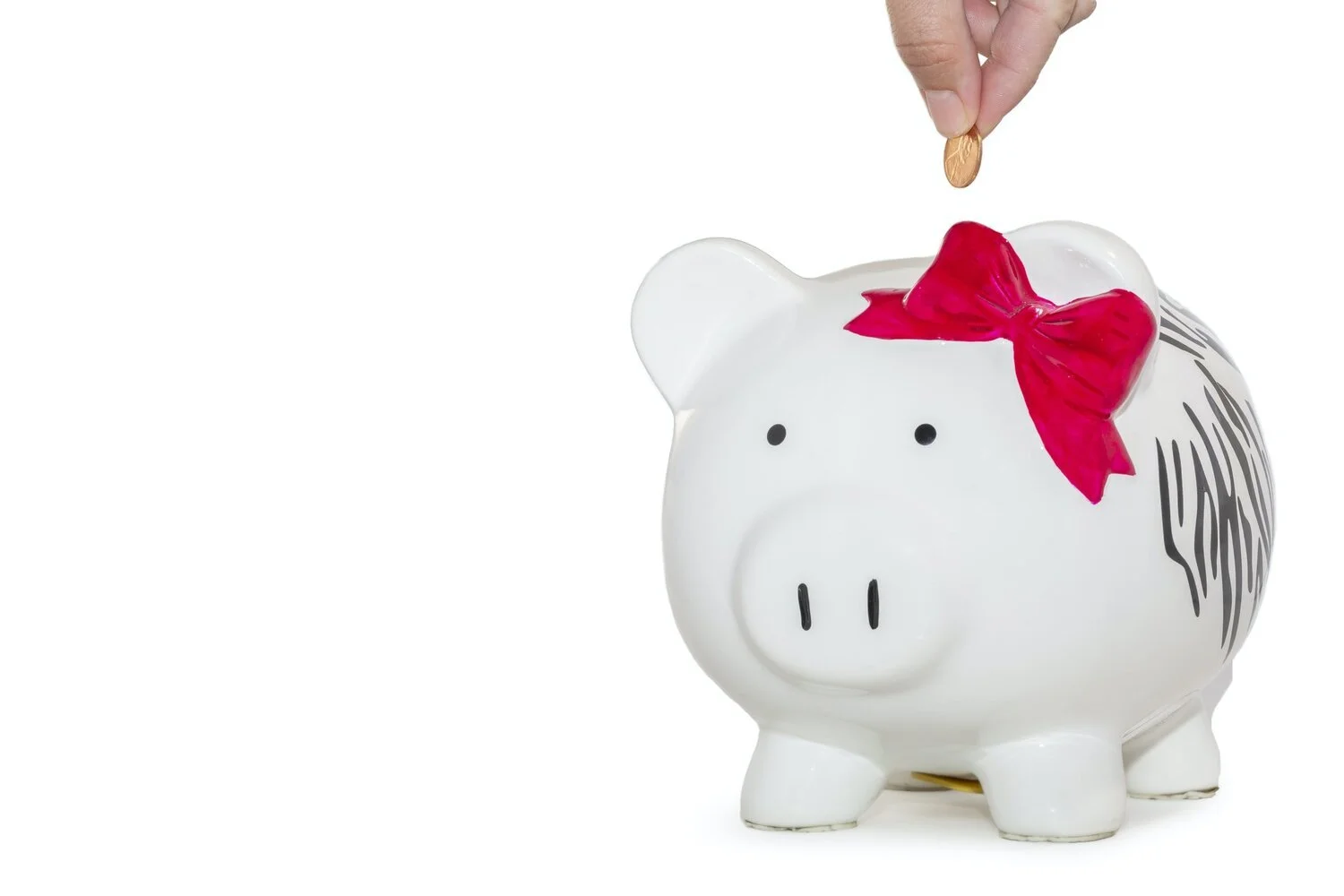 Hand dropping a coin into a white piggy bank with a red bow on its head and black and white zebra pattern on its side, against a white background.