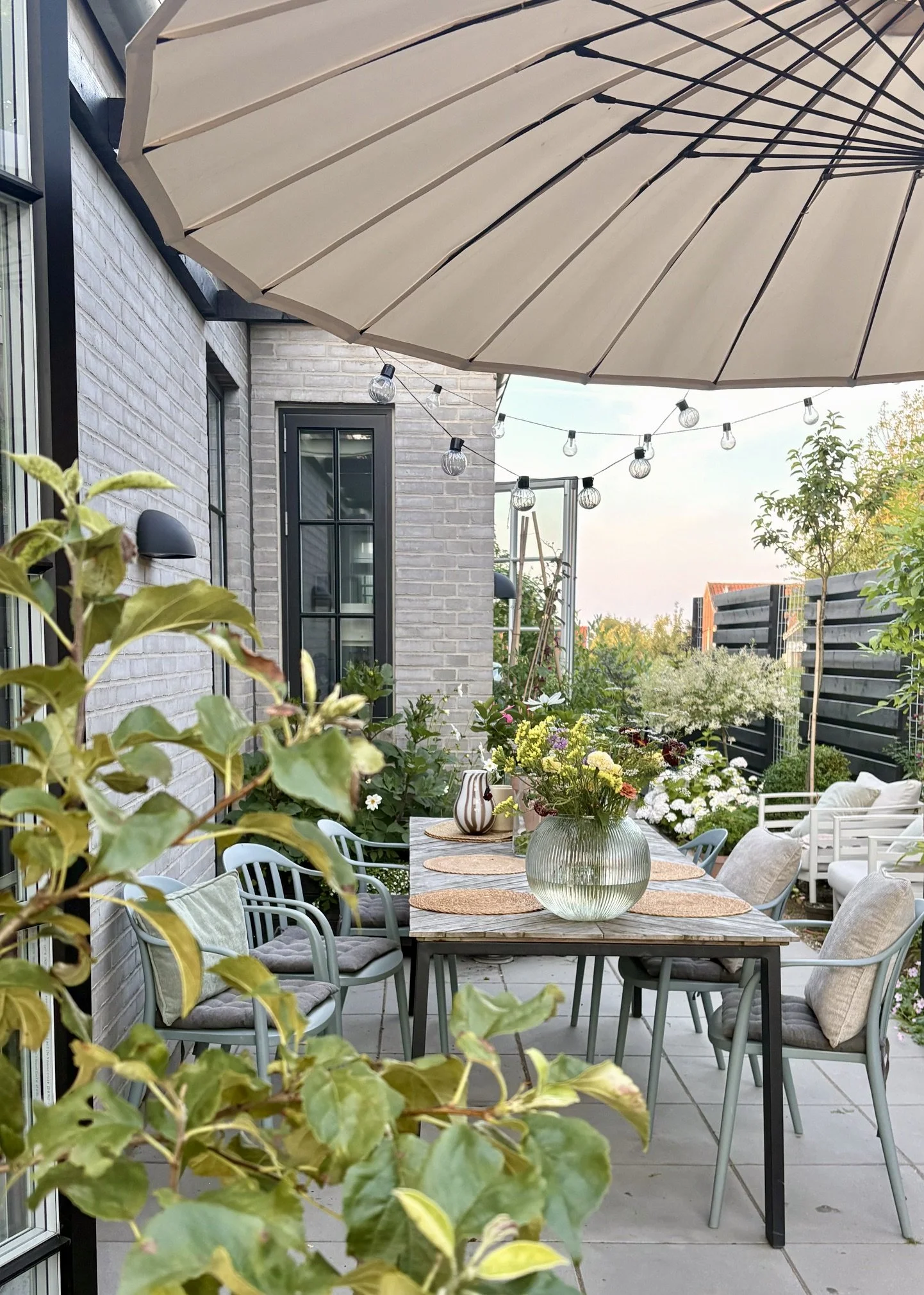 Lille skandinavisk terrasse med hyggeligt loungehjørne, hørpuder, urtepotter og klatreplanter. Aftenlys og rolig stemning med naturlige materialer.