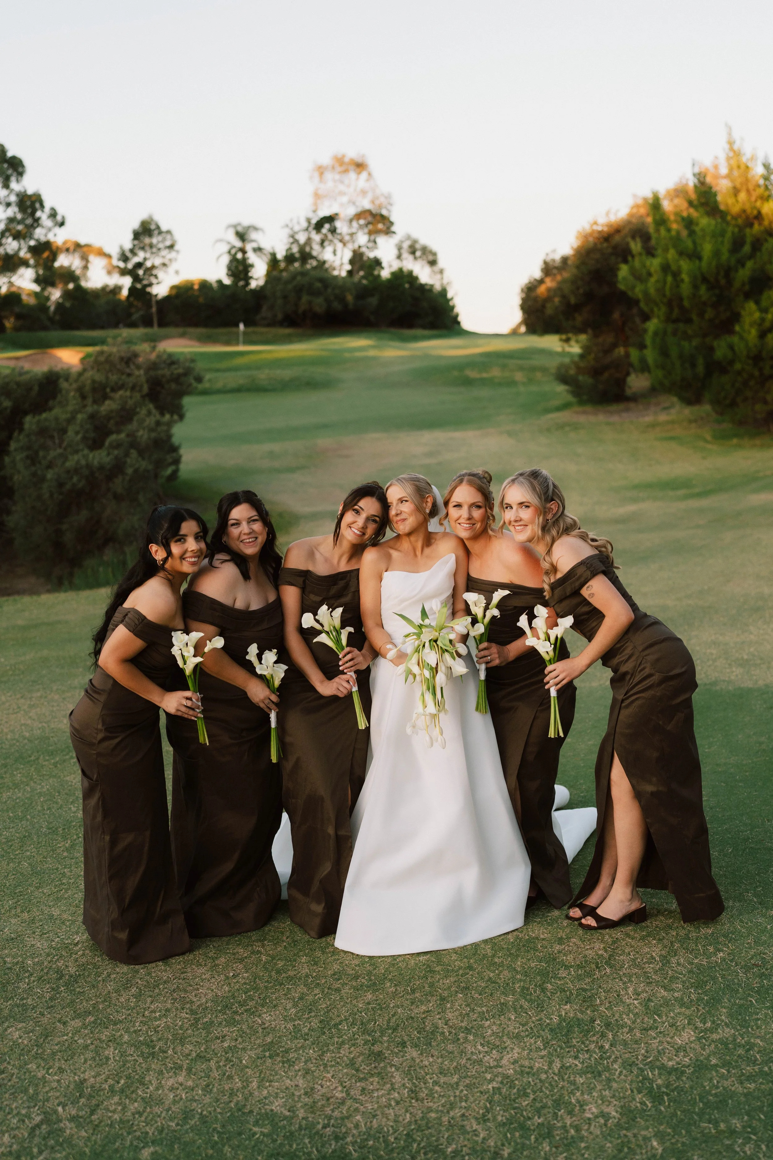 A group of six women, dressed in formal gowns, posing on a golf course with greenery and trees in the background, holding white calla lily bouquets, during sunset.