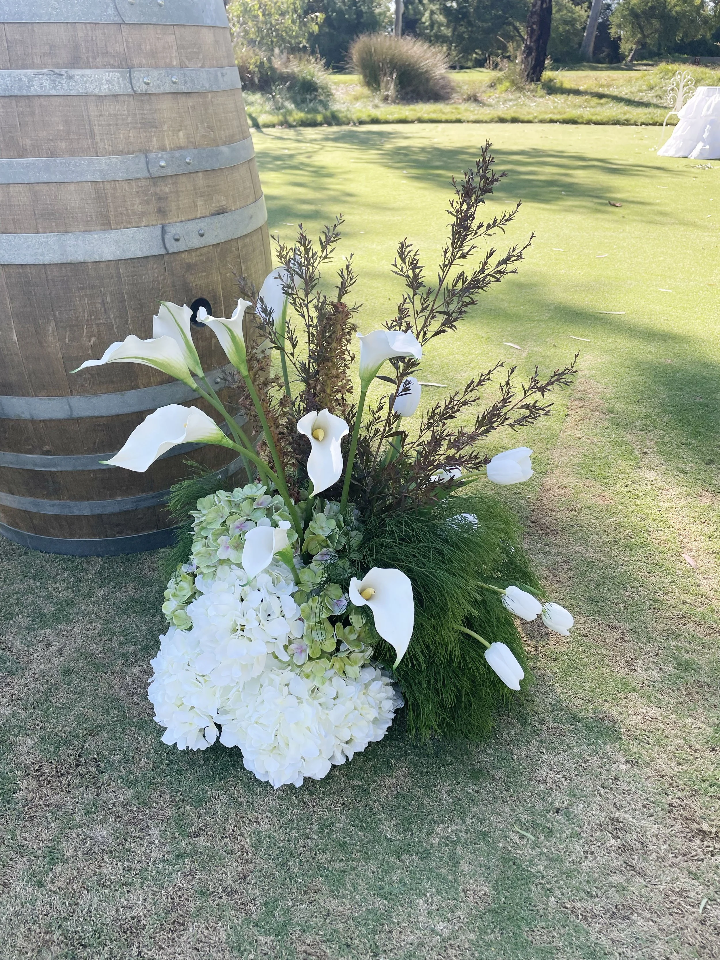 Arrangement of white calla lilies, hydrangeas, and foliage placed next to a wooden barrel outdoors on a grassy area with trees and bushes in the background.