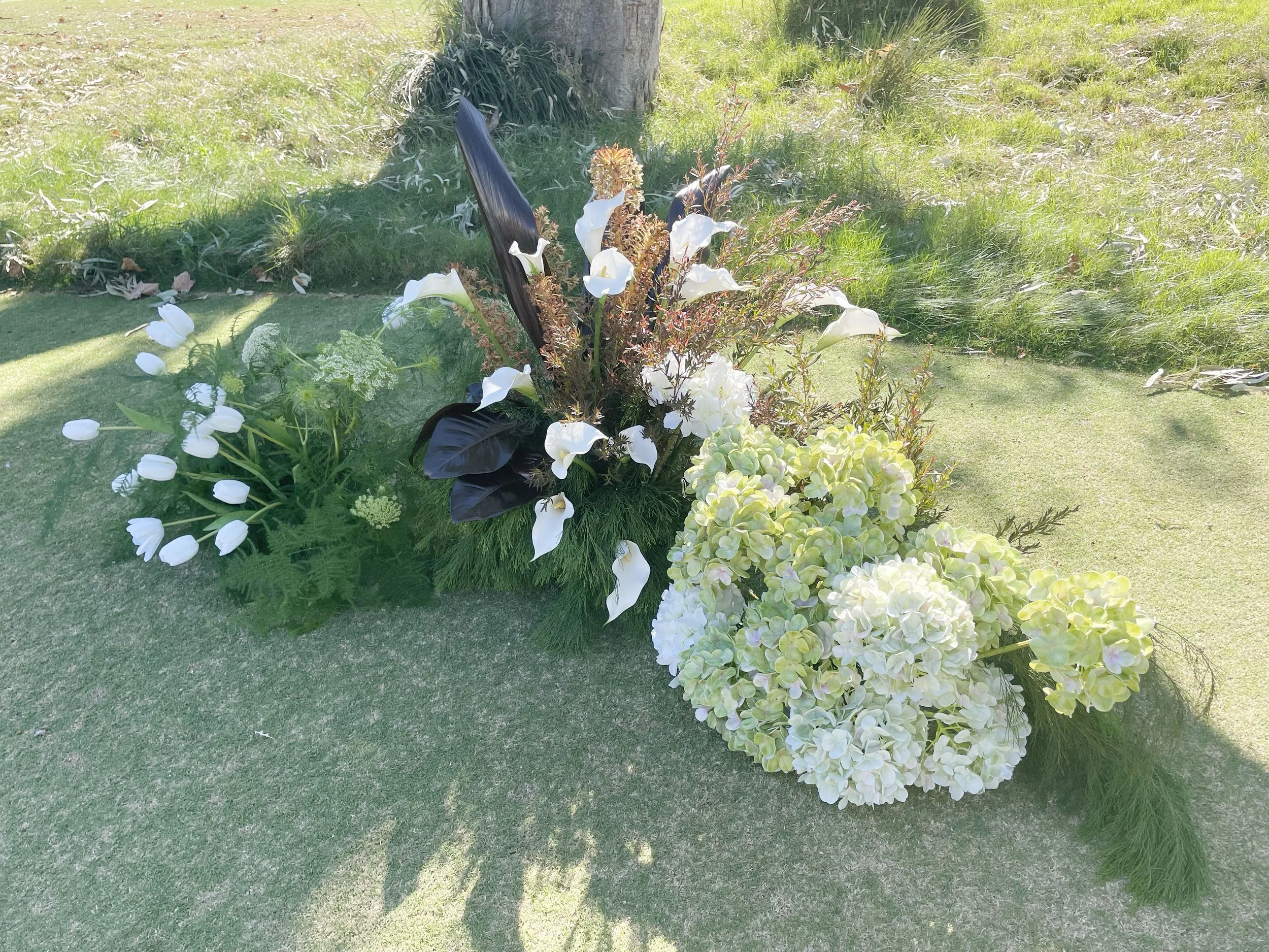 A floral arrangement with white calla lilies, green hydrangeas, and dark purple leaves, placed on a lawn with grass and a tree in the background.