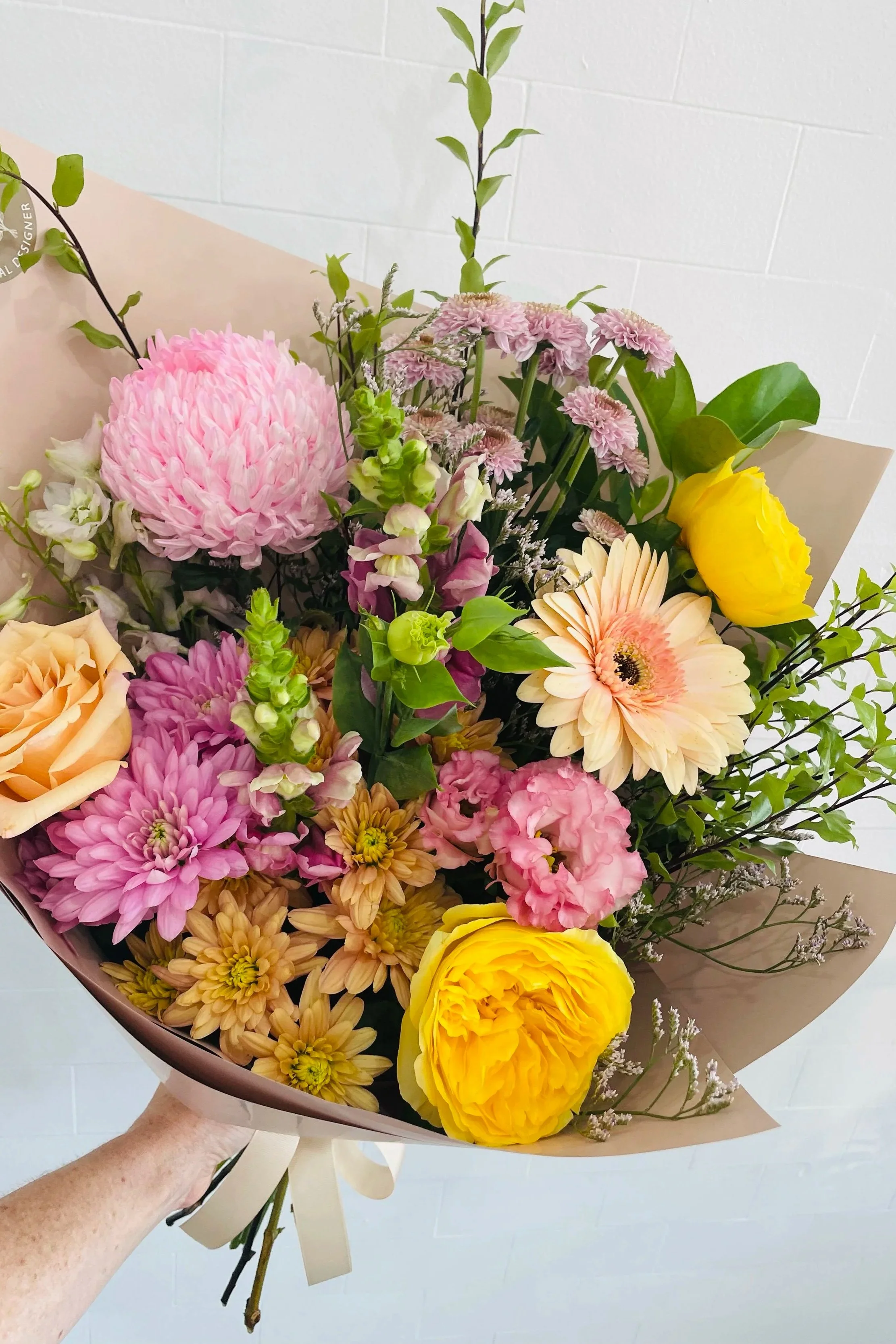 A hand holding a bouquet of mixed flowers, including pink chrysanthemums, yellow roses, and light pink gerbera daisies, wrapped in brown paper with a cream-colored ribbon.
