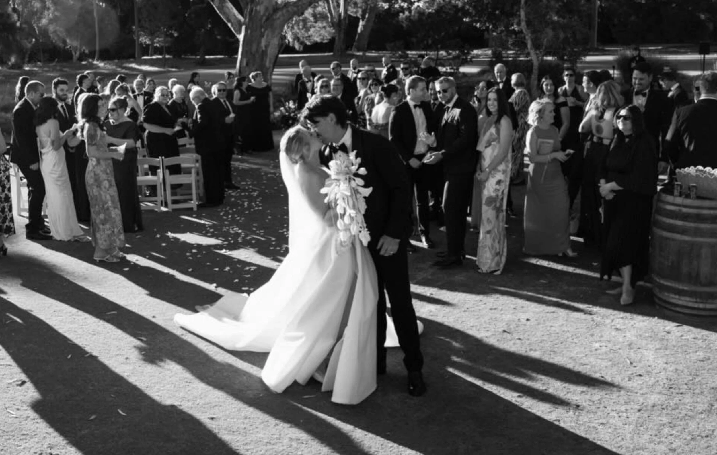 A black and white photo of a wedding scene outdoors with a bride and groom walking together, surrounded by guests dressed in formal attire.
