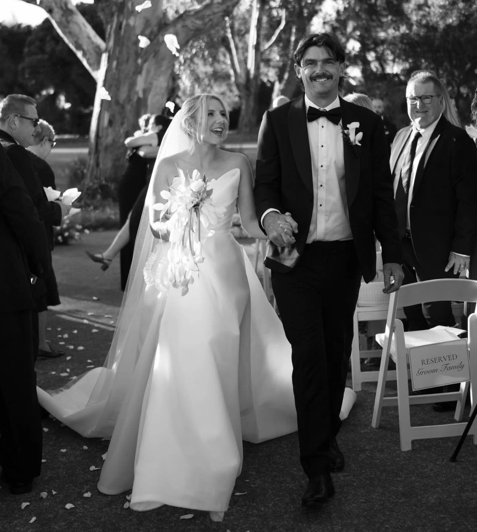 Black and white photo of a bride and groom walking hand in hand at their outdoor wedding, surrounded by guests, with a reserved sign on a chair in the foreground.