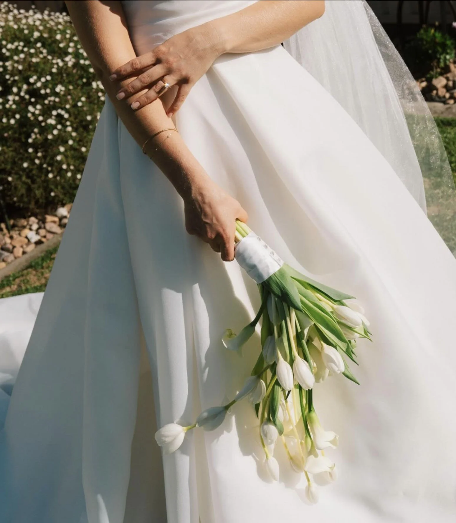 A woman in a white dress holding a bouquet of white calla lilies with her left hand, while her right arm is crossed over her chest, standing outdoors in a garden setting.
