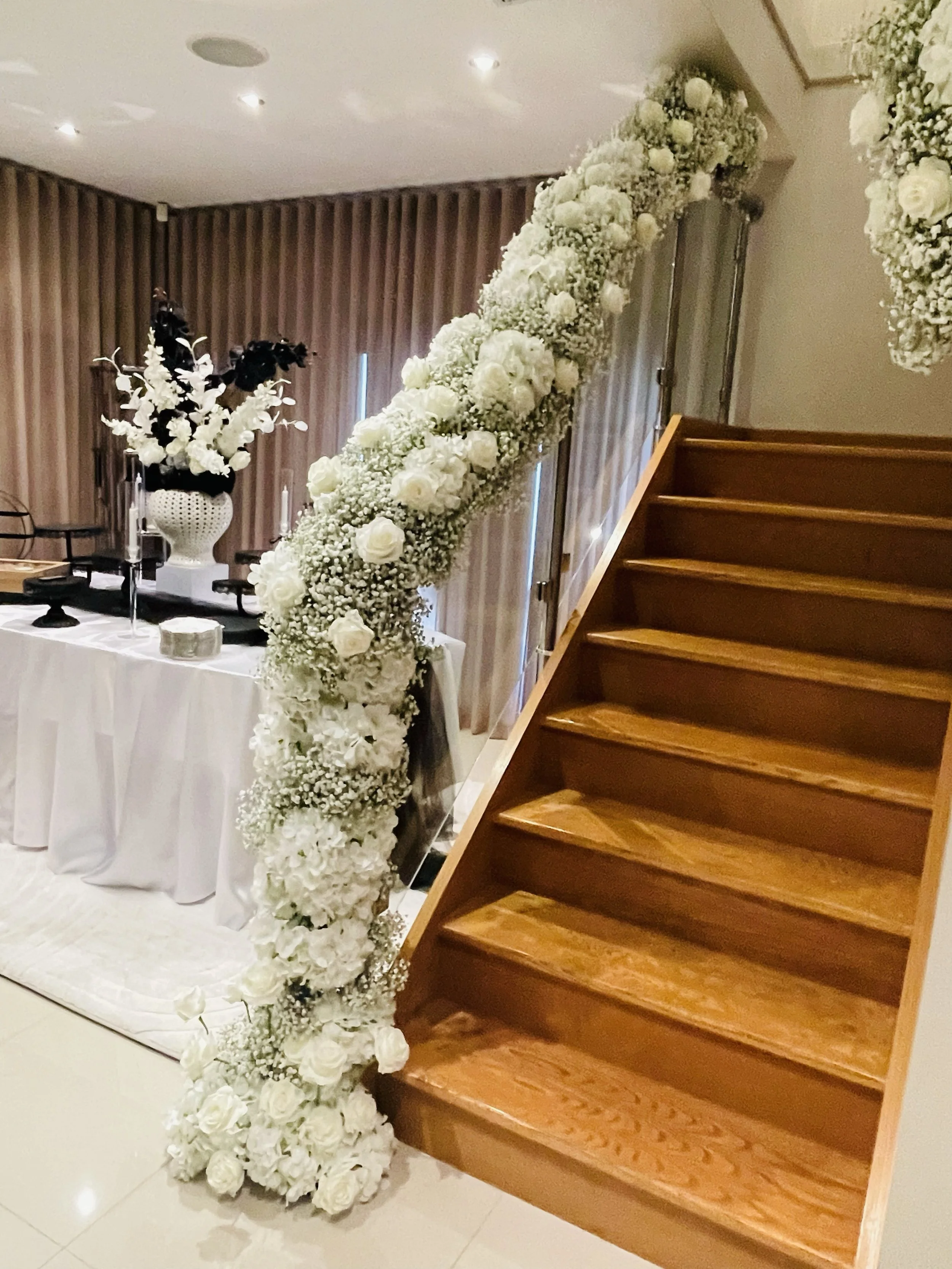 A floral arrangement made of white roses, baby's breath, and various white flowers on a staircase and table in a decorated event space.