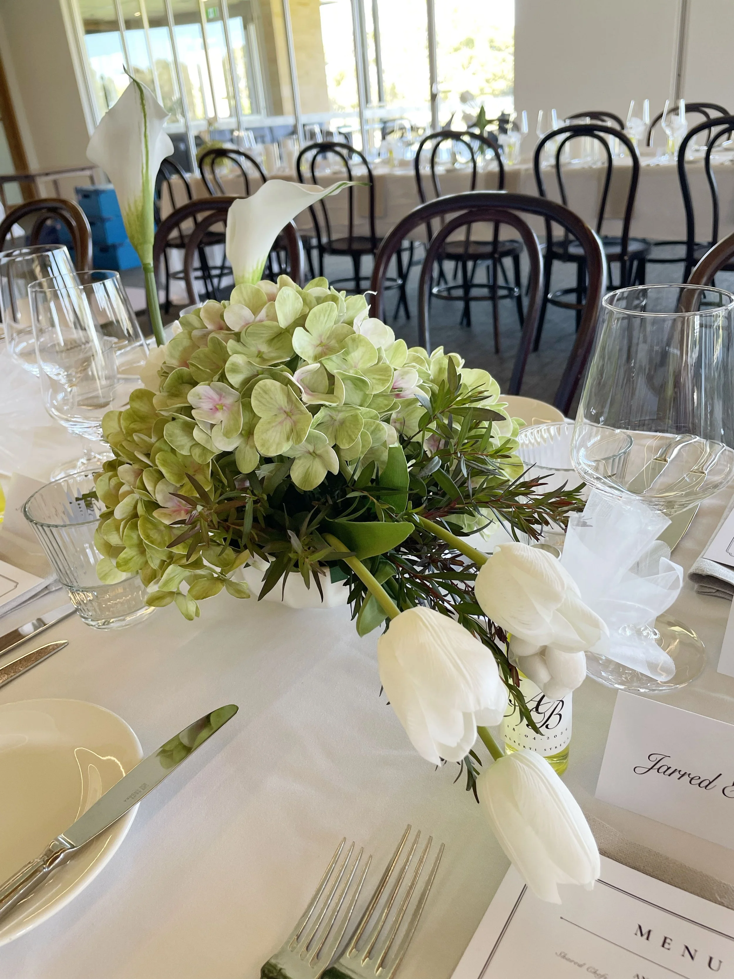 A floral centerpiece with white calla lilies and green hydrangeas on a white tablecloth, set for a formal event, with champagne glasses and silverware arranged around it.