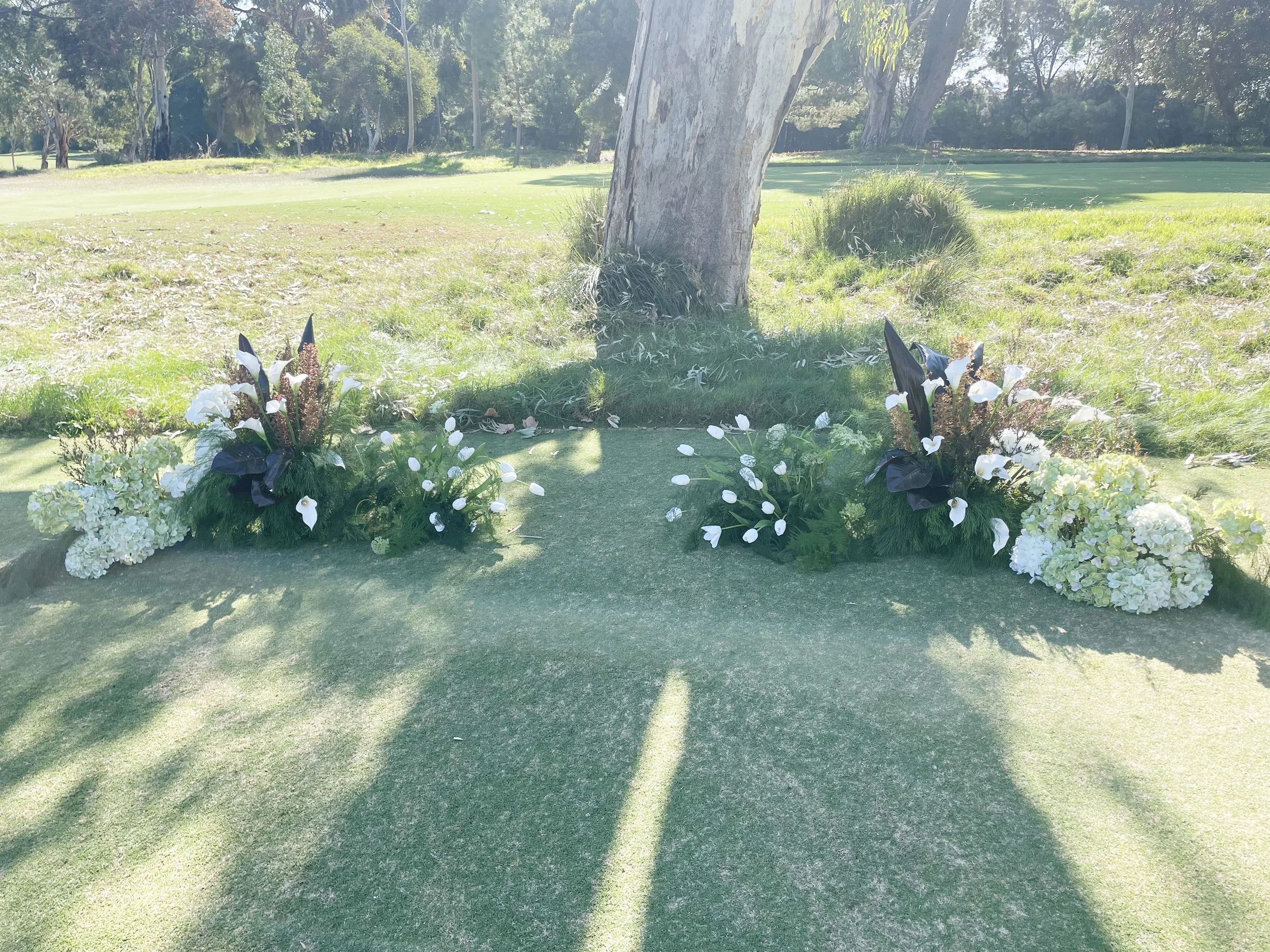 Flowers arranged on a patch of grass with a large tree trunk behind them, in a park-like setting with trees and a golf course in the background.