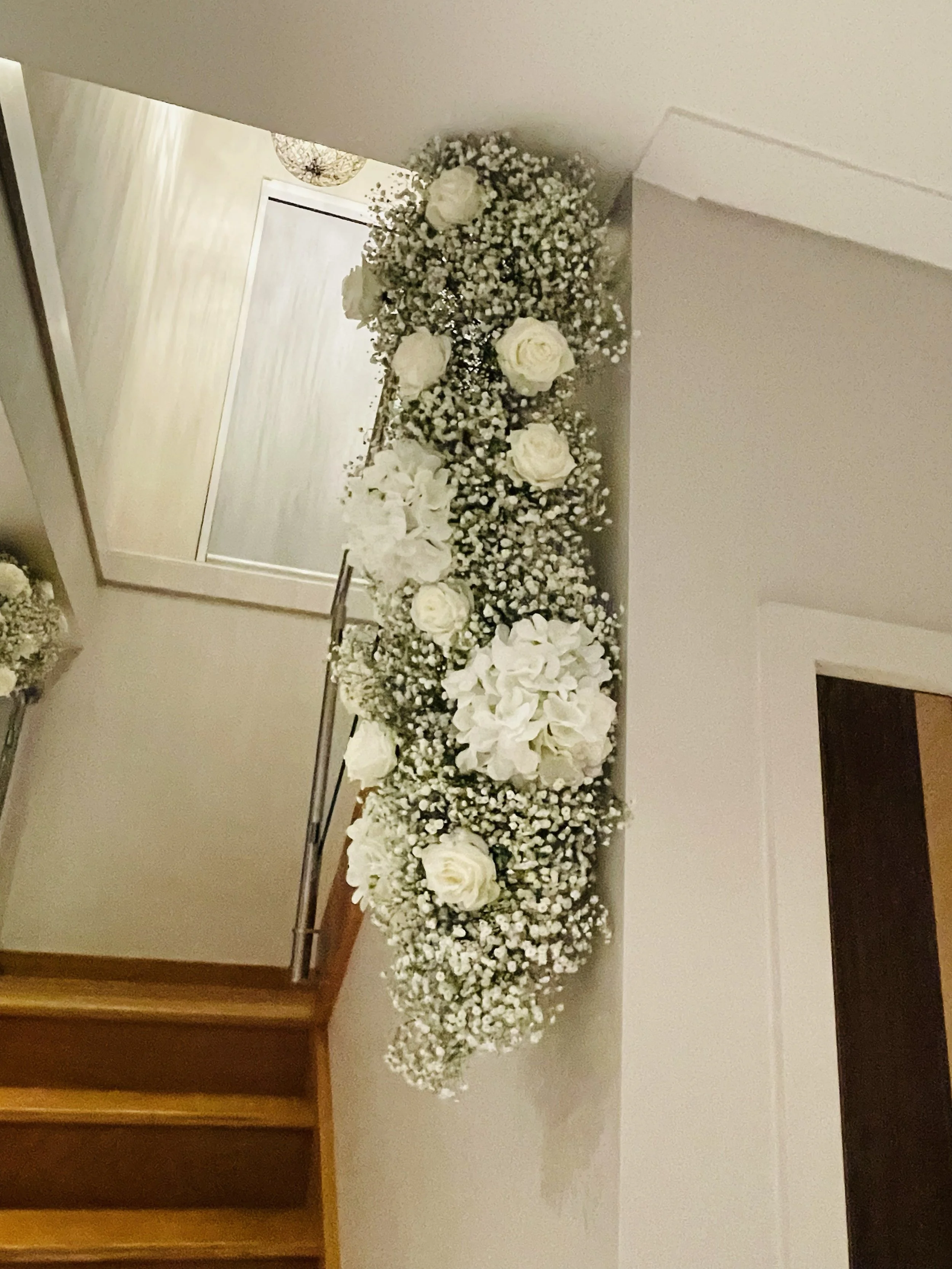 White floral arrangement with roses and baby's breath on a table in front of a mirror in a doorway.