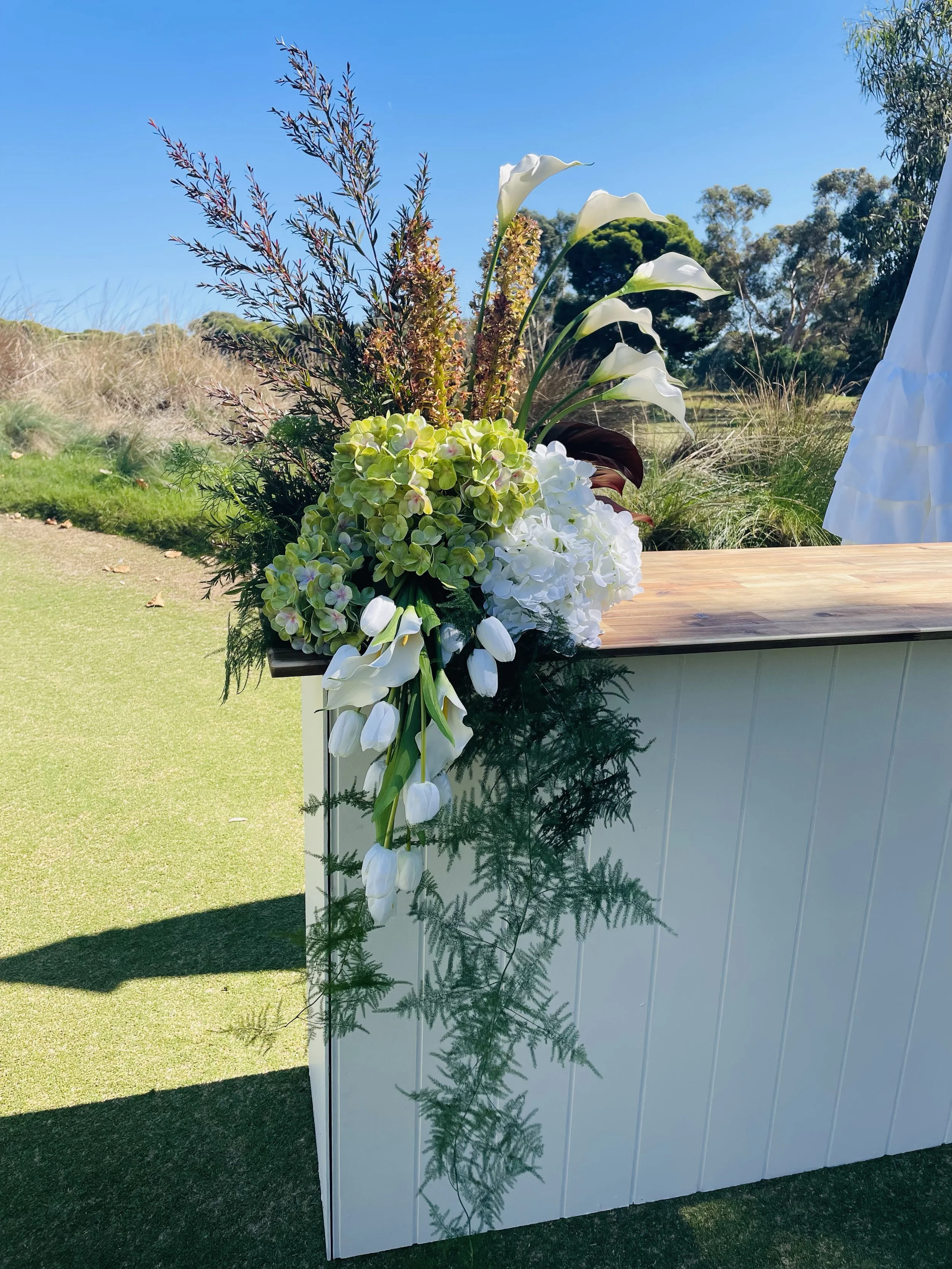 A floral arrangement with white calla lilies, green hydrangeas, and various foliage, placed on a wooden surface outdoors under a blue sky with trees in the background.