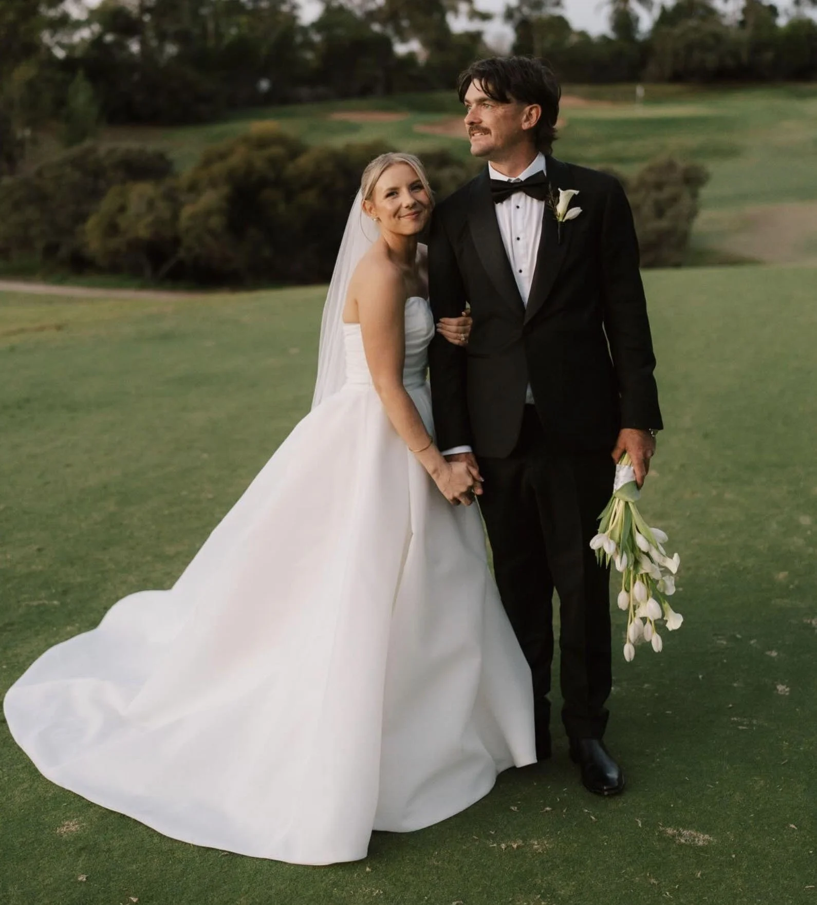 A bride and groom standing on a grassy field during a wedding, holding hands, with the bride smiling and the groom holding a bouquet of white flowers.