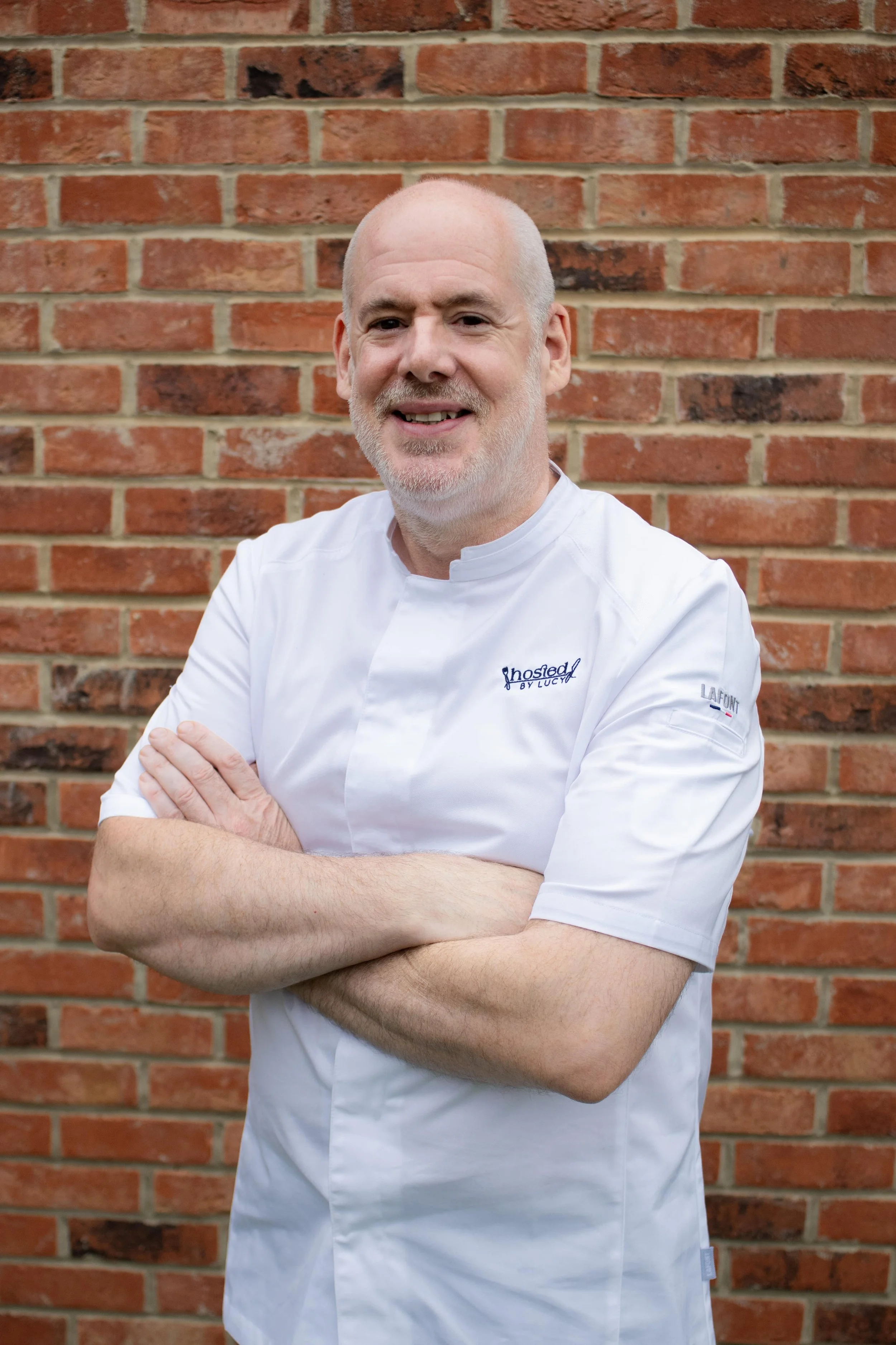 A middle-aged man with a beard and bald head stands with arms crossed in front of a brick wall, wearing a white chef's coat with the logo 'Hostad by Lucy' on the chest.