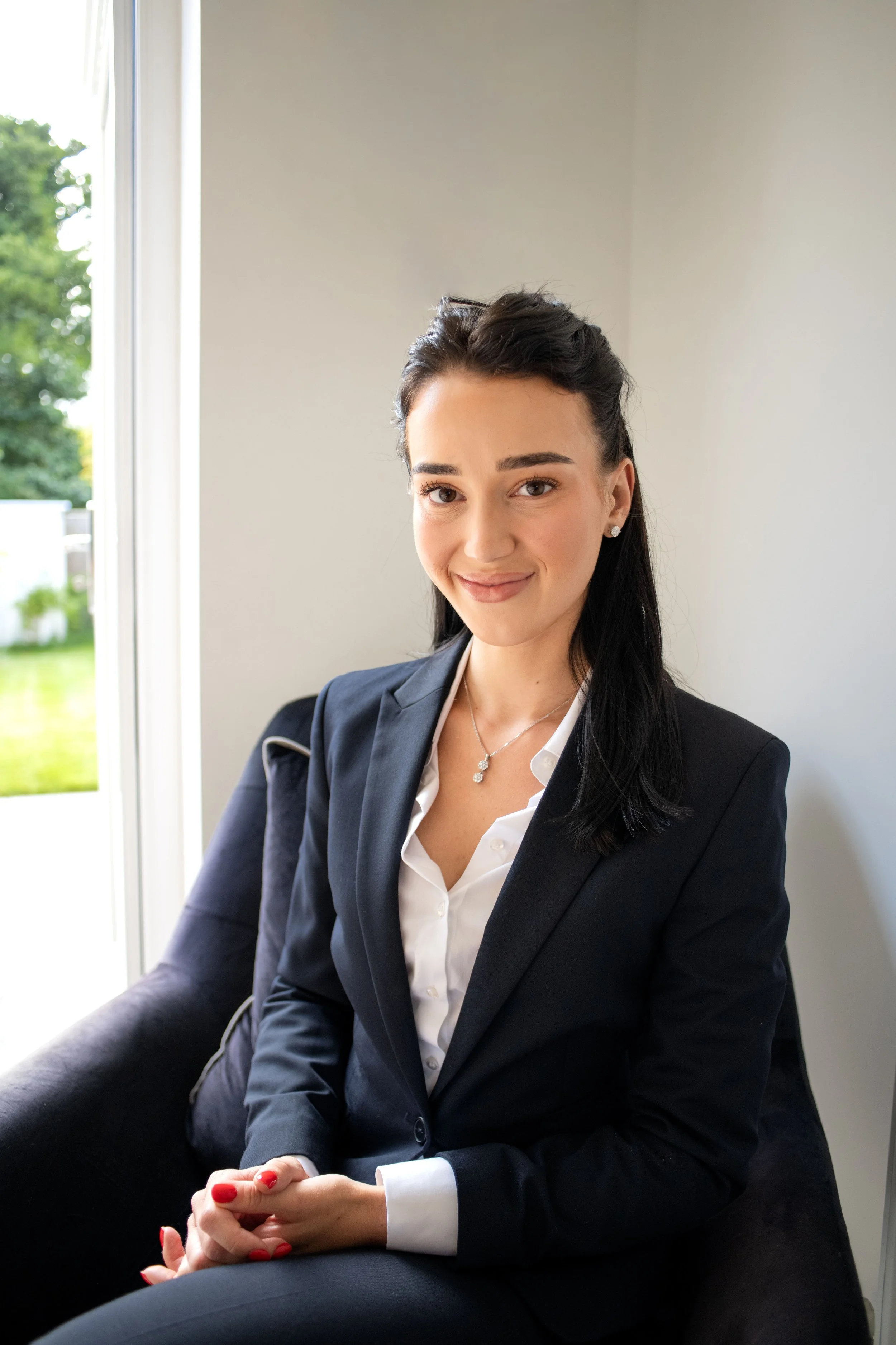 A young woman in a black business suit and white blouse sitting on a dark couch in a bright room near a large window, smiling at the camera.