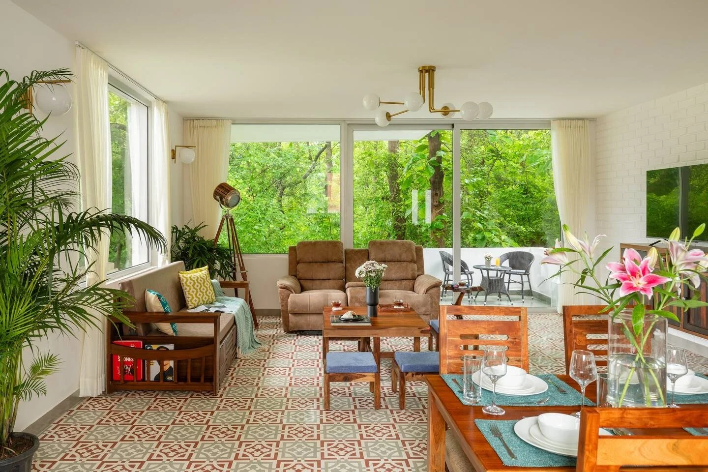 Bright living room with large windows showing green trees, decorated with plants, wooden furniture, a brown armchair, a beige couch, a glass-topped coffee table, a dining table set with plates, glasses, and flowers, and a balcony with outdoor seating.