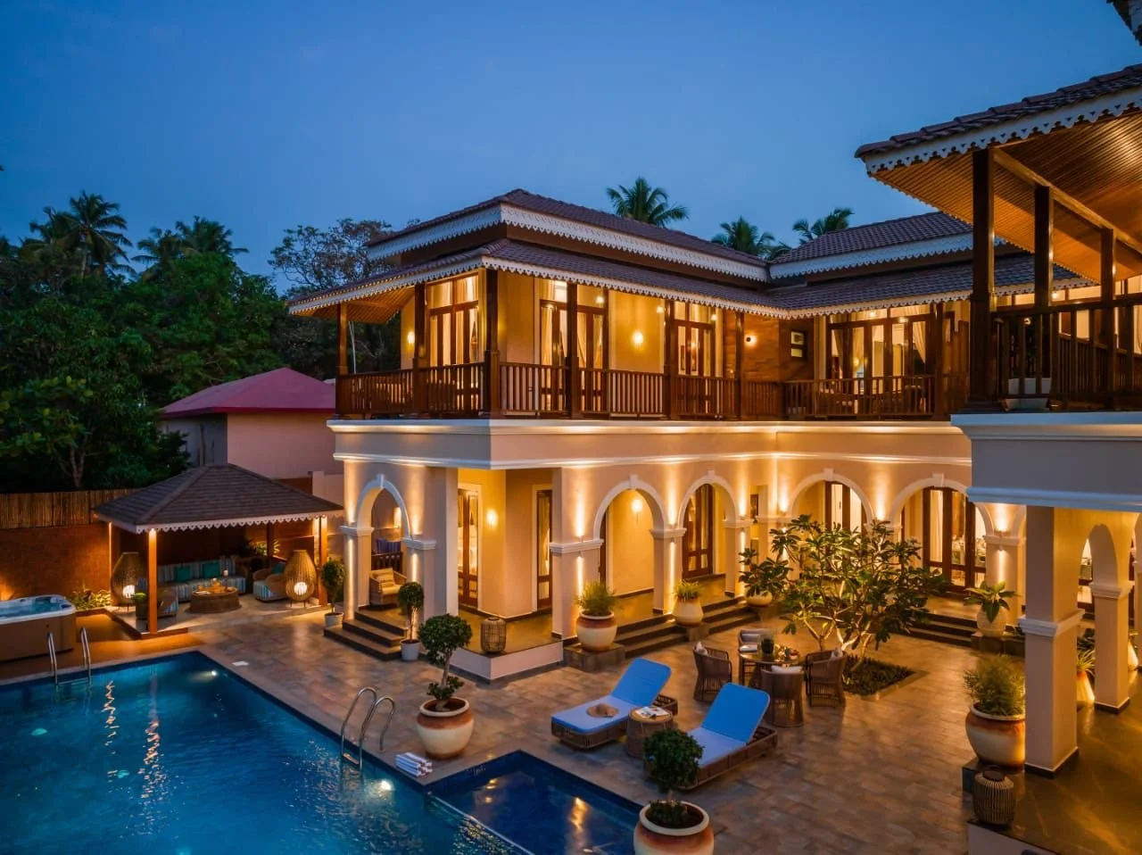 Luxury two-story house with warm lighting, balcony, swimming pool, outdoor seating, palm trees, and tropical landscaping at dusk.
