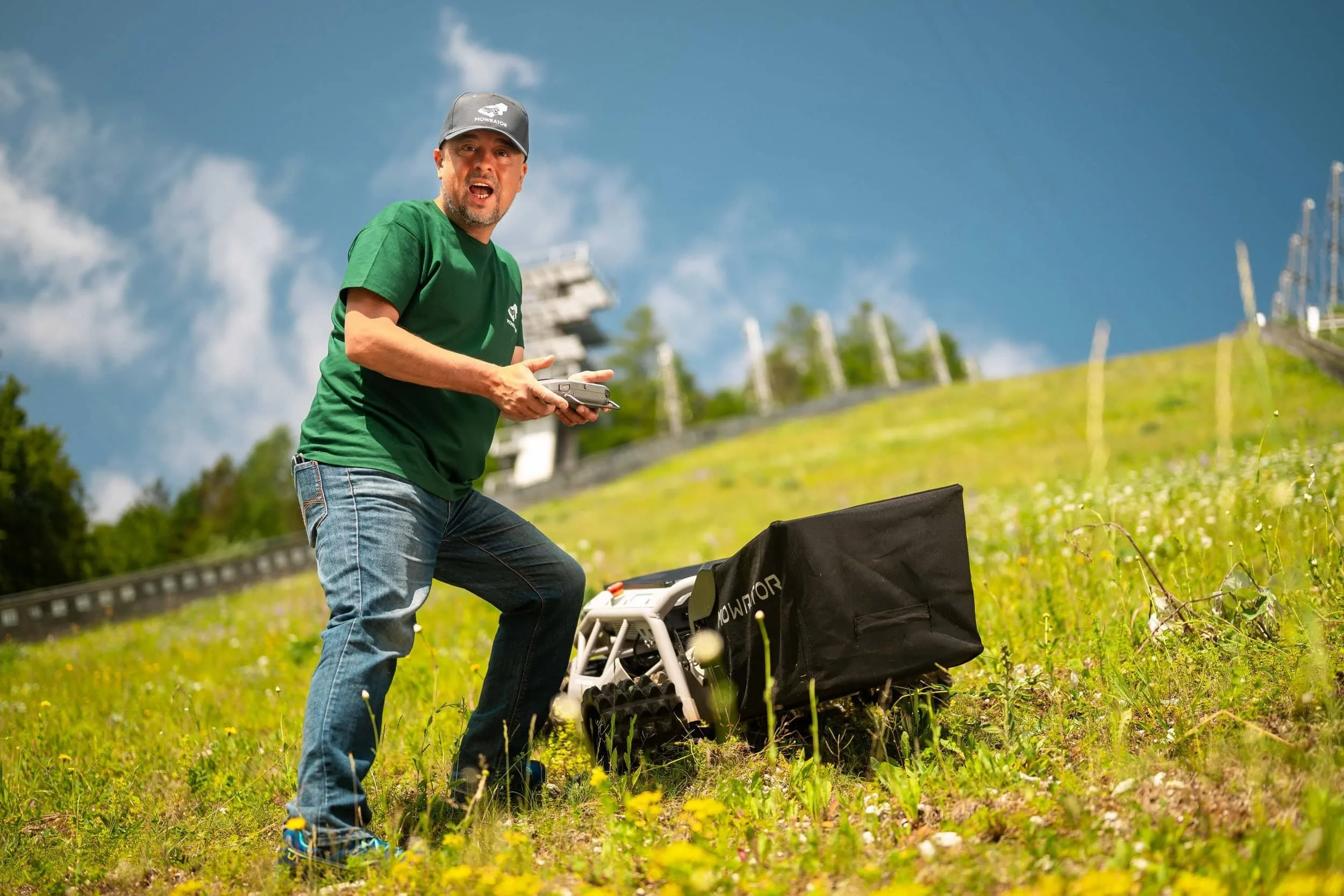 Man operating a drone outdoors on a grassy hill, holding a remote control with a drone on the ground nearby.