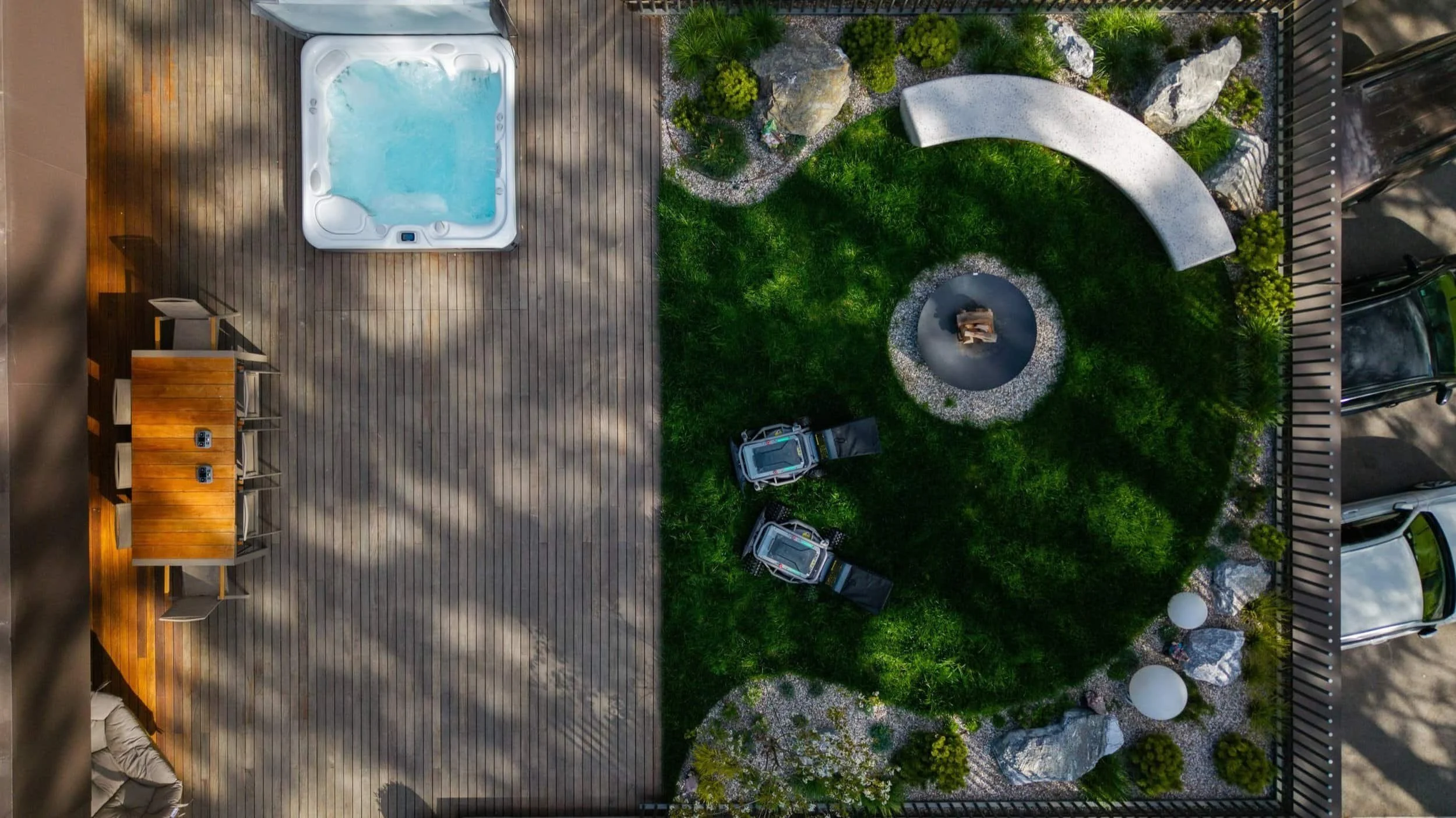 An aerial view of a backyard with a hot tub on a wooden deck on the left, a grassy area with a fire pit at the center, surrounded by rocks and plants, and a section of parking with cars on the right.