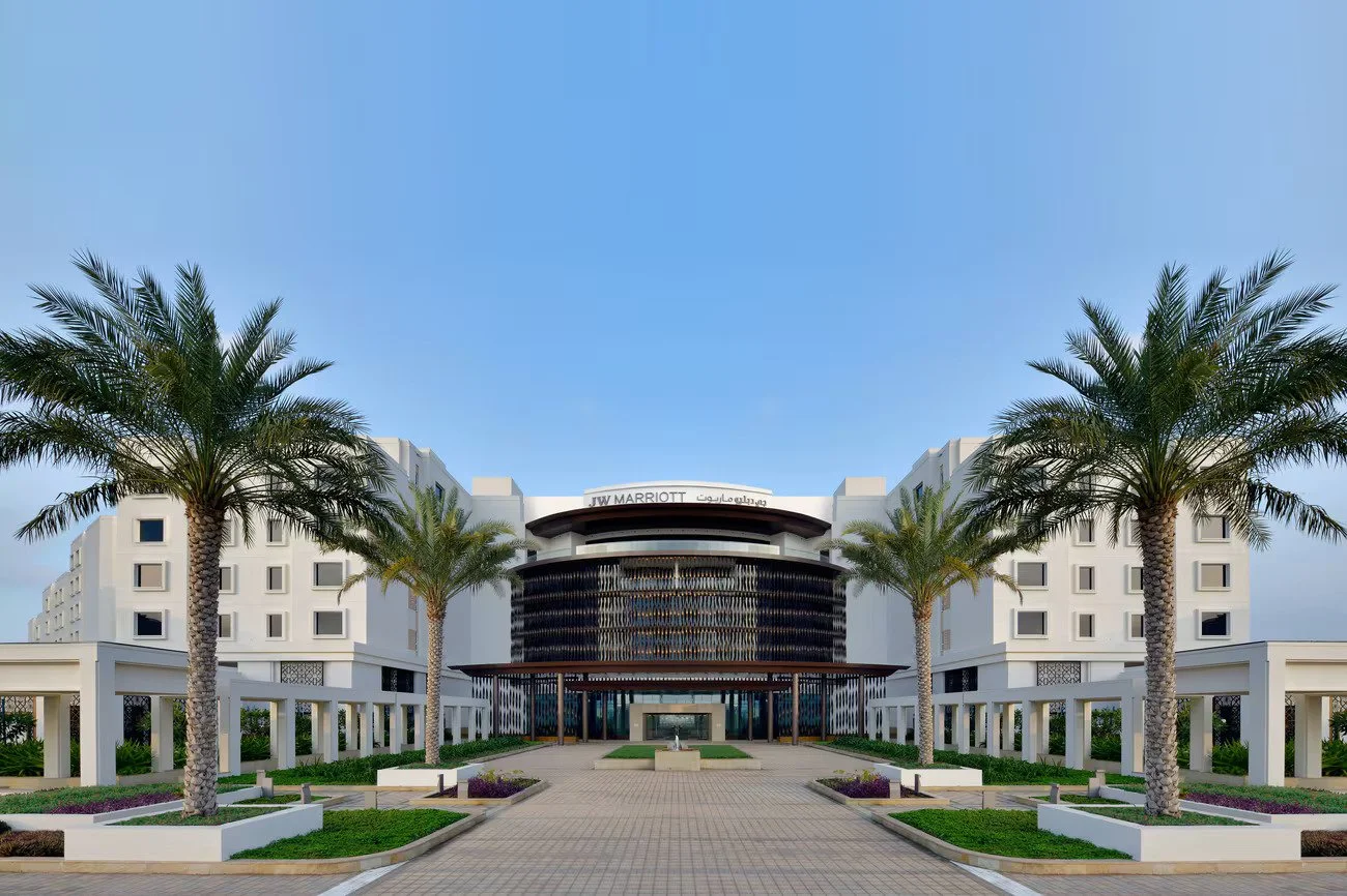 Modern JW Marriott hotel with white exterior, palm trees lining a paved walkway, and a clear blue sky.