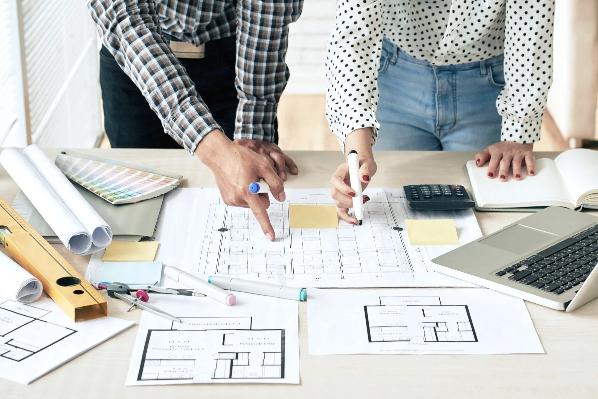 Two people working on architectural blueprints and floor plans at a desk with tools, a laptop, and notes.