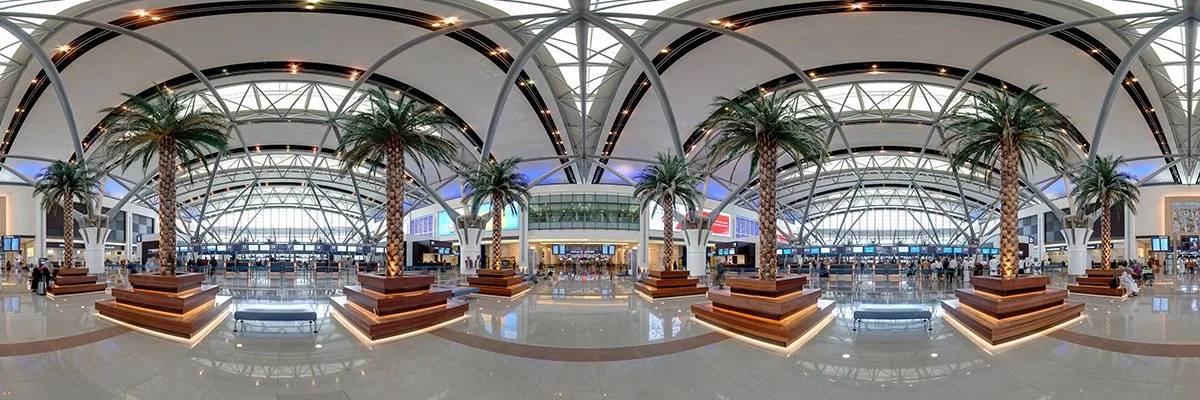 Interior of an airport terminal with palm trees, seating areas, and check-in counters.