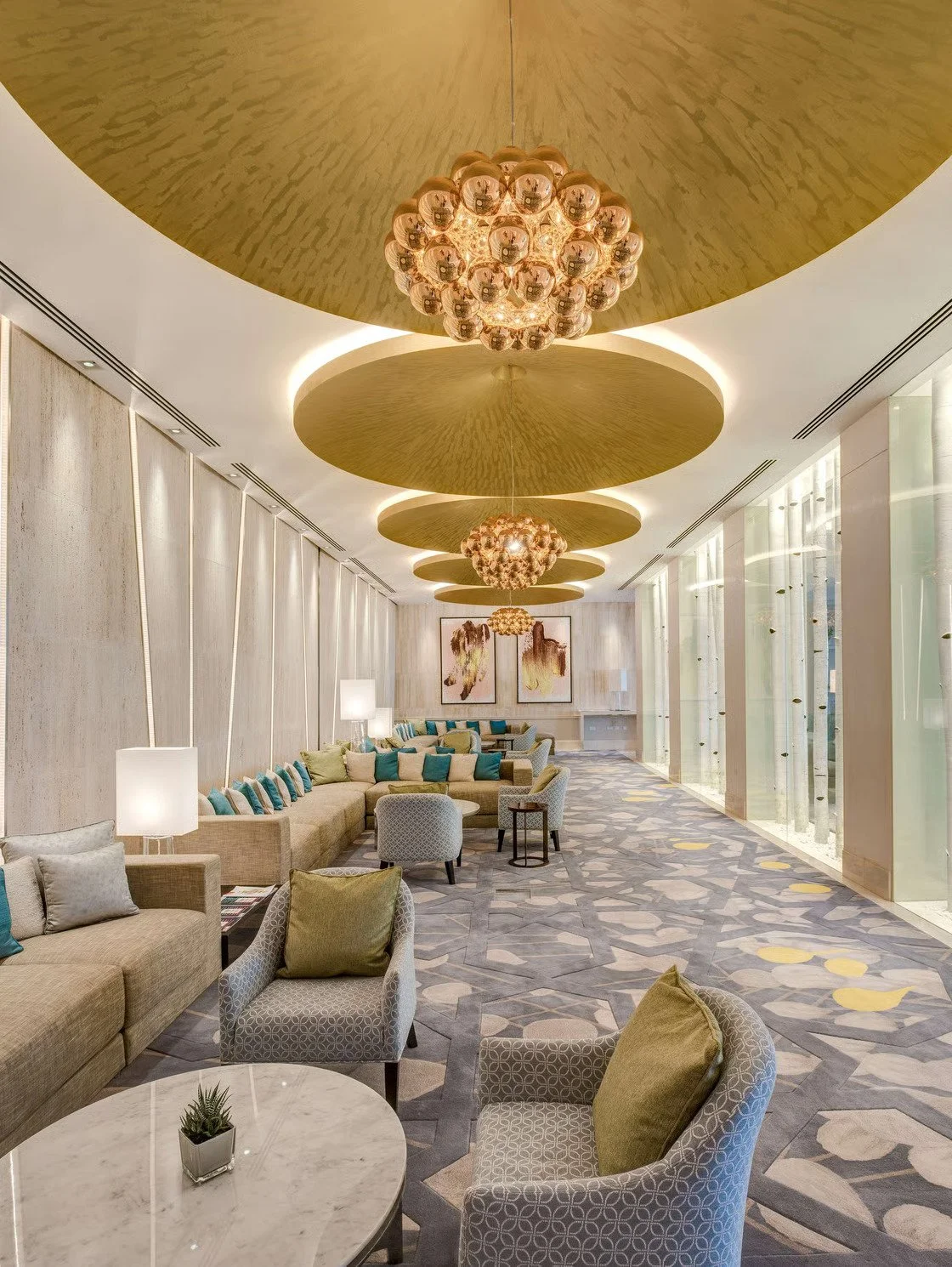 Luxurious hotel lounge with modern decor, grey patterned armchairs, sofas with teal and beige cushions, marble coffee table, tall lamps, gold ceiling features, ornate gold hanging chandeliers, abstract artwork, large windows with decorative glass panels, patterned carpet.