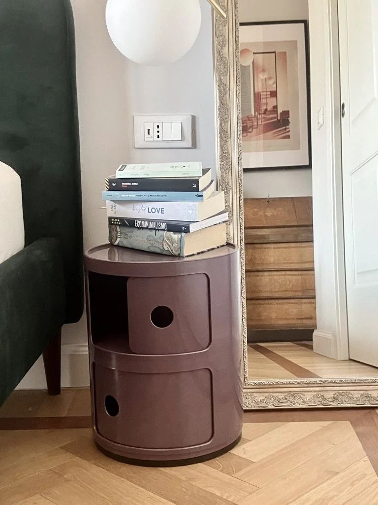 Tavolino da notte rotondo color rosa con tre libri impilati sopra, uno di questi si intitola 'Economie Ministeriale'. C'è anche una grande cornice con specchio e una lampada bianca sopra. La stanza ha pareti bianche e pavimento in legno.