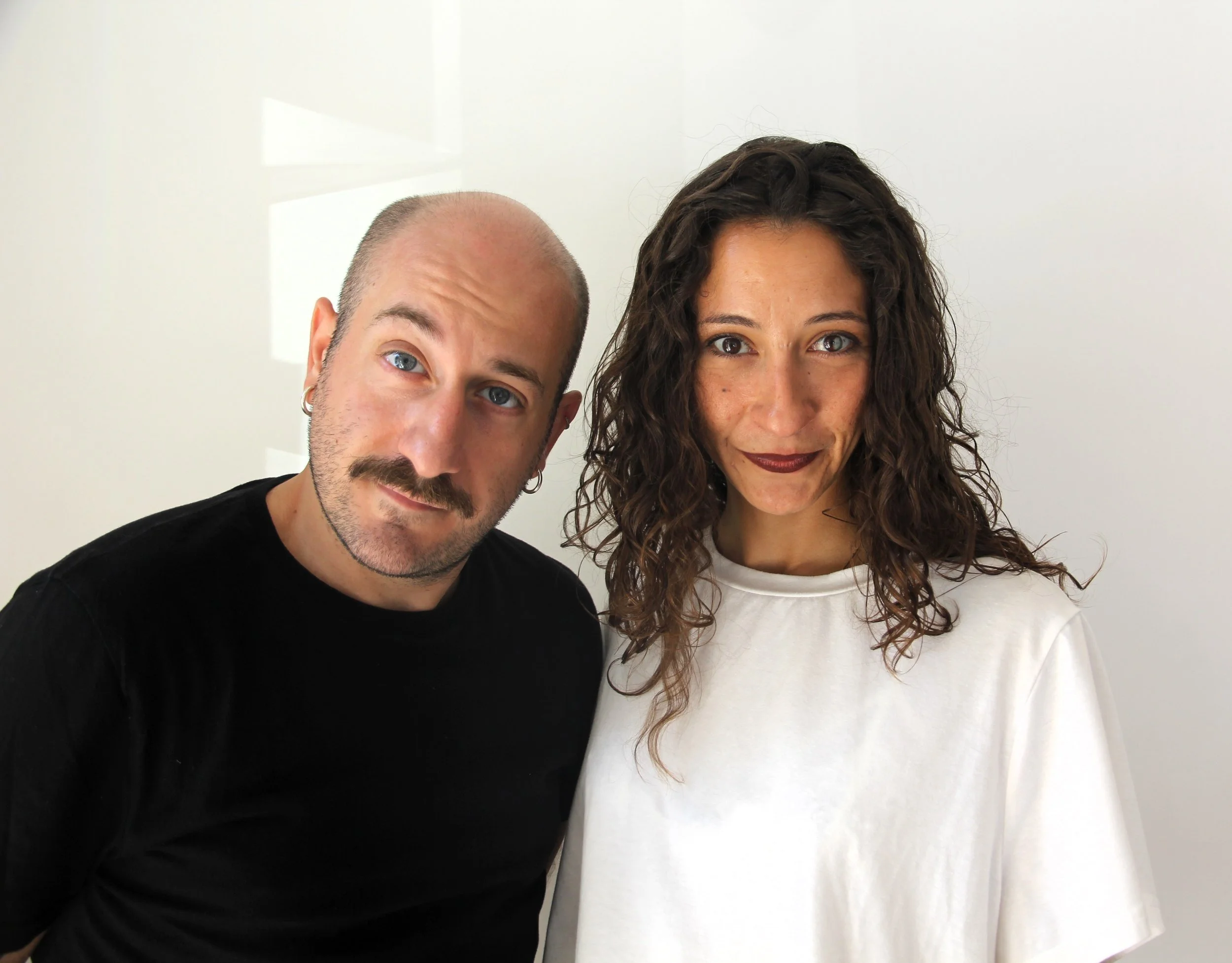 Una coppia di giovani posa per una foto, lui con una maglietta nera, lei con una maglietta bianca, entrambi sorridenti e in un ambiente neutro con pareti chiare.