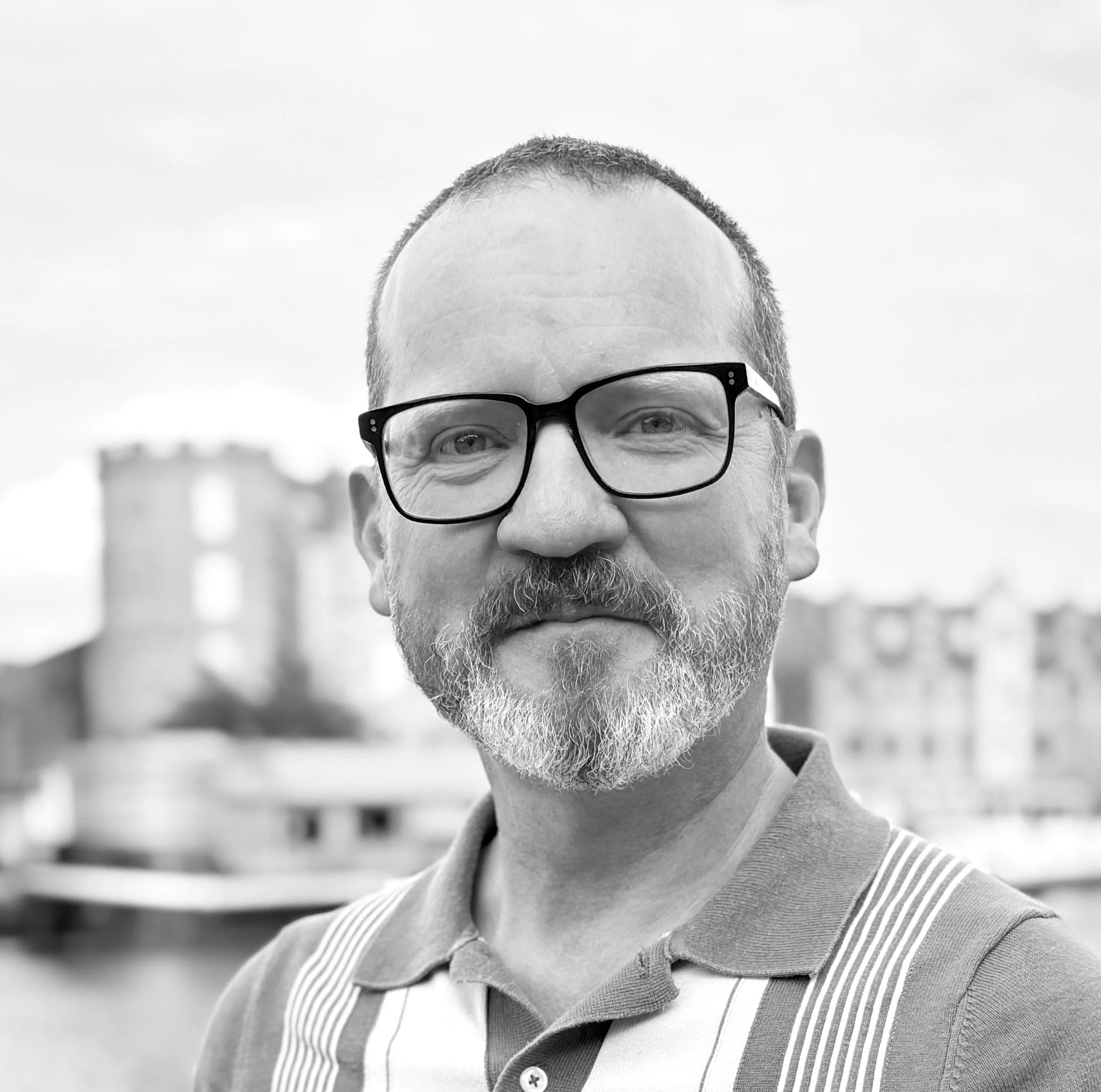 A black-and-white photo of a middle-aged man with glasses, a beard, and short hair, standing outdoors with blurred buildings and a tower in the background.