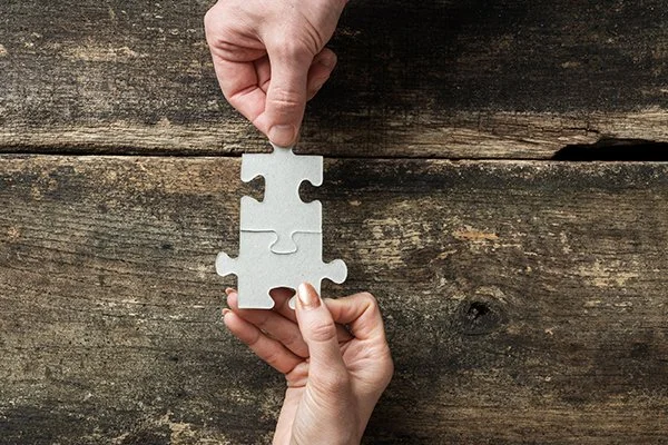 Accredited BABCP and EMDR UK Psychotherapist specialising in kink friendly, BDSM, fetish related trauma and self esteem issues. Offering cognitive behavioural therapy (CBT) - Two hands assembling a puzzle piece over a wooden surface.