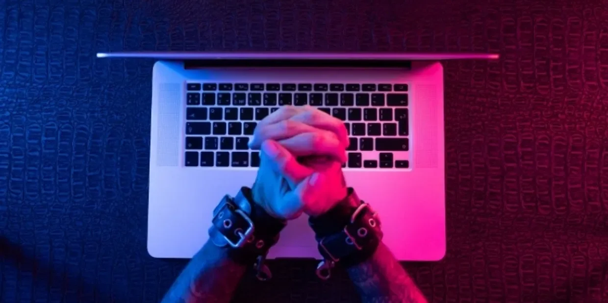 Accredited BABCP and EMDR UK Psychotherapist specialising in kink friendly, BDSM, fetish related trauma and self esteem issues. Person with chained wrists praying over a laptop with blue and pink neon lighting.