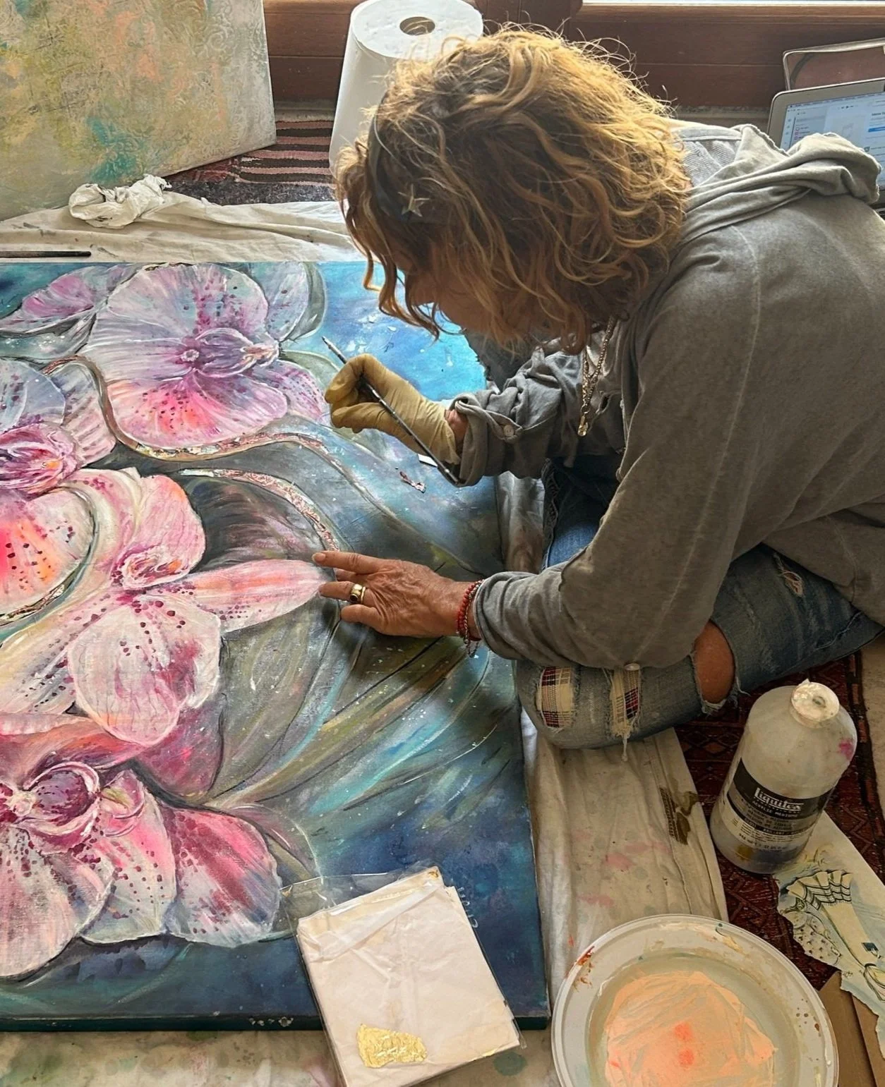An artist painting large pink and purple orchids on a canvas, sitting on the floor surrounded by painting supplies and a laptop.
