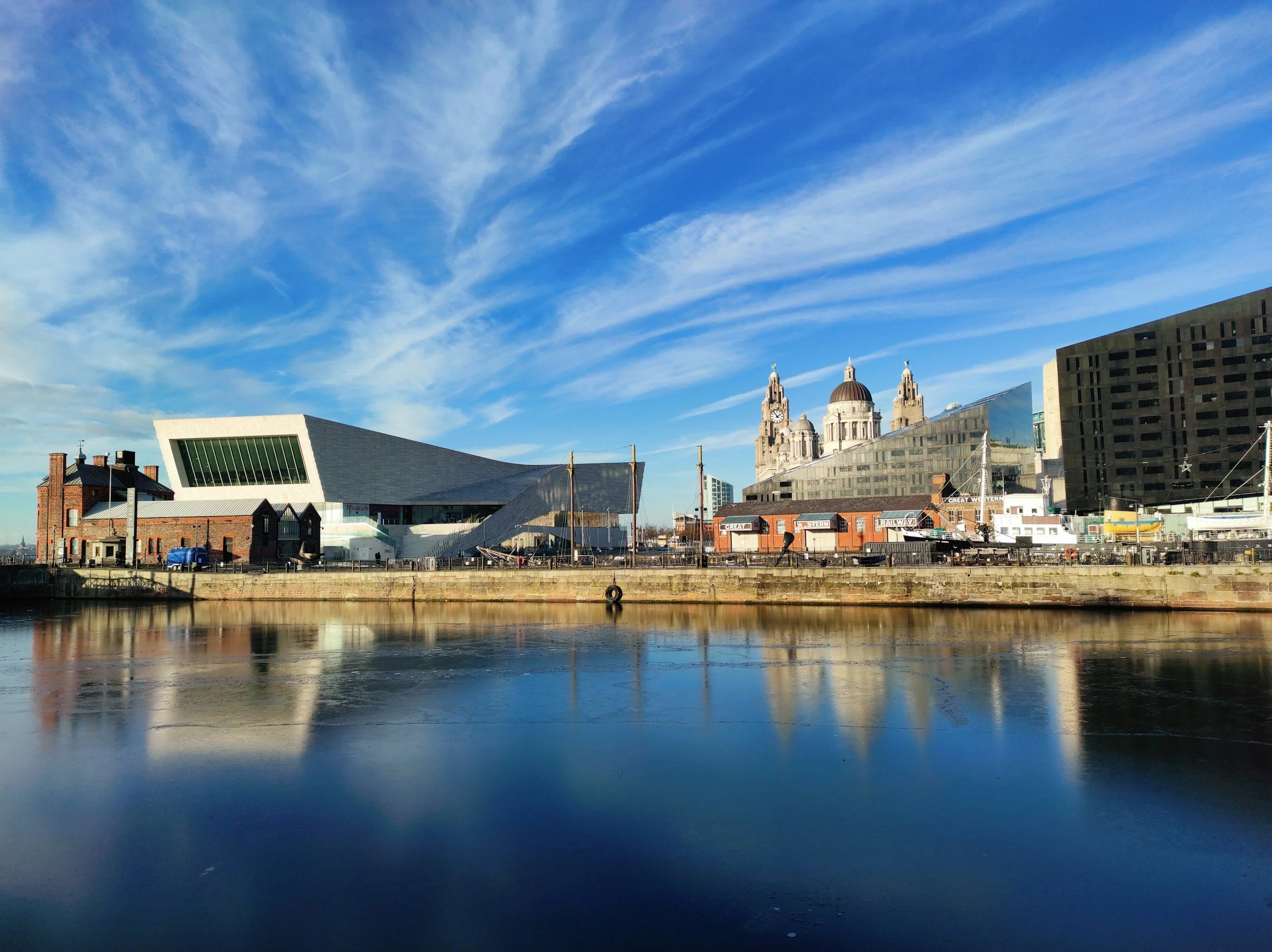 Skyline view of a city with modern and historic buildings reflected on a body of water, under a mostly clear blue sky with wispy clouds.