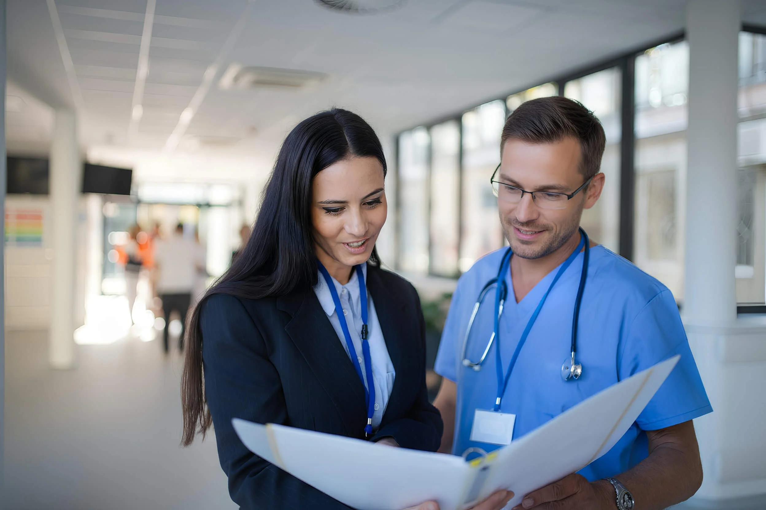 A female doctor and a male nurse reviewing medical documents together in a hospital corridor.