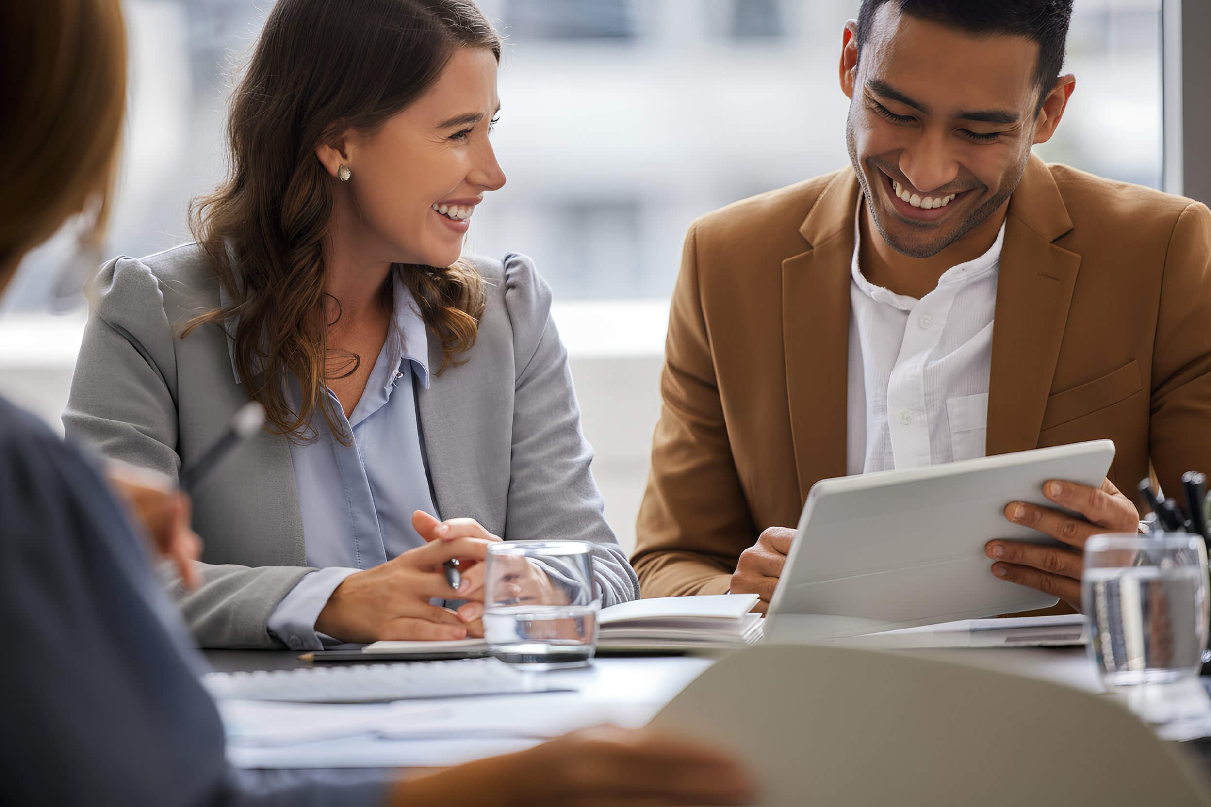 Business meeting in a modern office with diverse colleagues. A woman smiles while looking at a man who is showing her something on a tablet. Others are in the background. The table has notebooks, glasses of water, and a pen.
