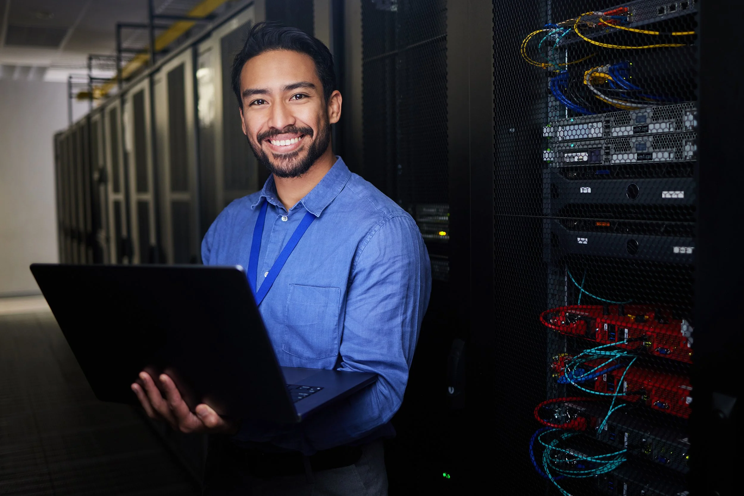 A smiling technician or IT professional in a blue shirt holding a laptop while standing next to server racks with network cables in a data center.