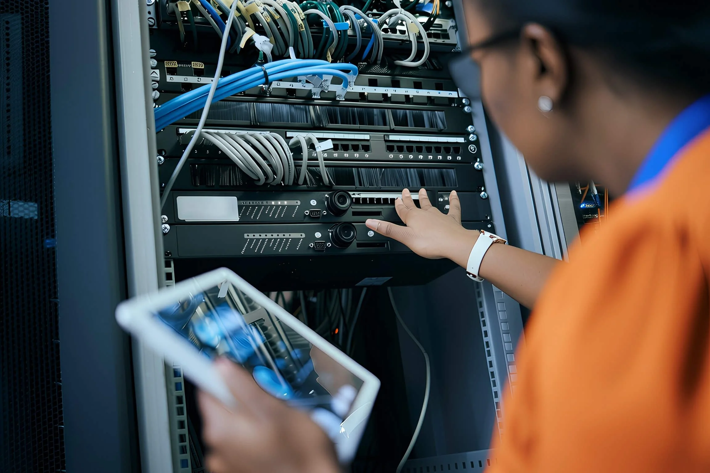 A person working on a server rack, connecting or disconnecting Ethernet cables. The person's face is partially visible, and they are wearing a wristband and earrings. The server rack contains multiple network cables and electronic components.