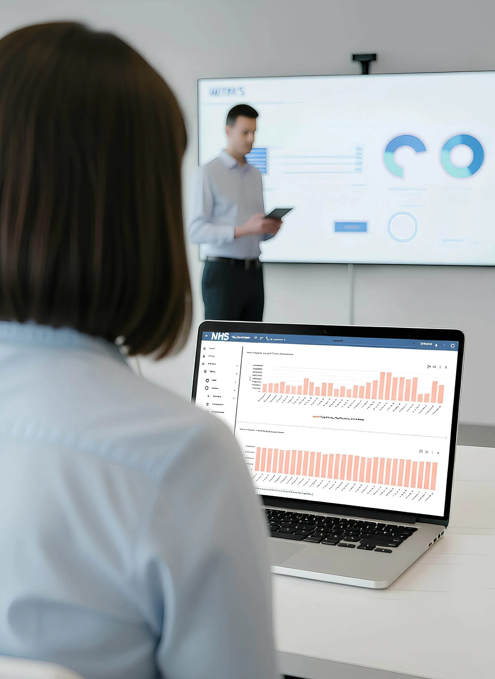 A woman is sitting at a desk with a laptop displaying a bar chart, and a man in a white shirt is standing in the background holding a smartphone in front of a large screen with various infographics.