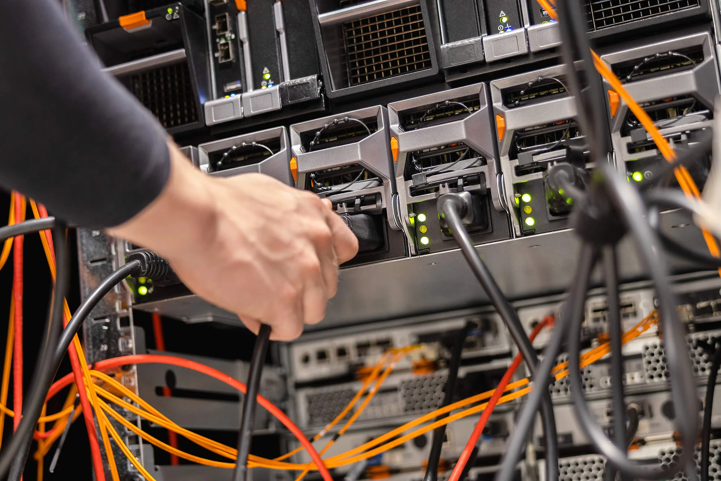 Close-up of a person's hand working on a server rack with multiple interconnected servers and numerous orange and black cables.