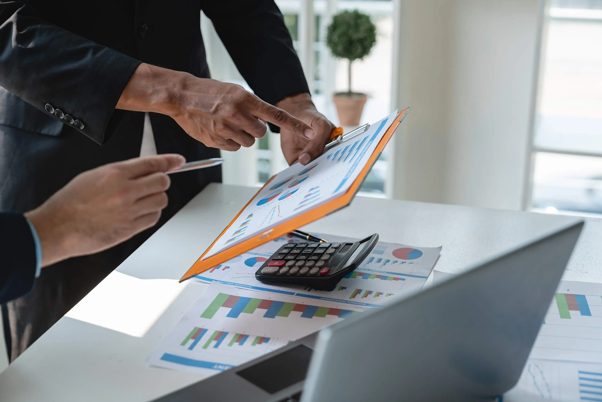 A person in a dark suit points at a document with blue and red graphical charts, while another person holds a pen. The desk has additional papers with graphs, a calculator, and a smartphone. Large windows in the background let in natural light.