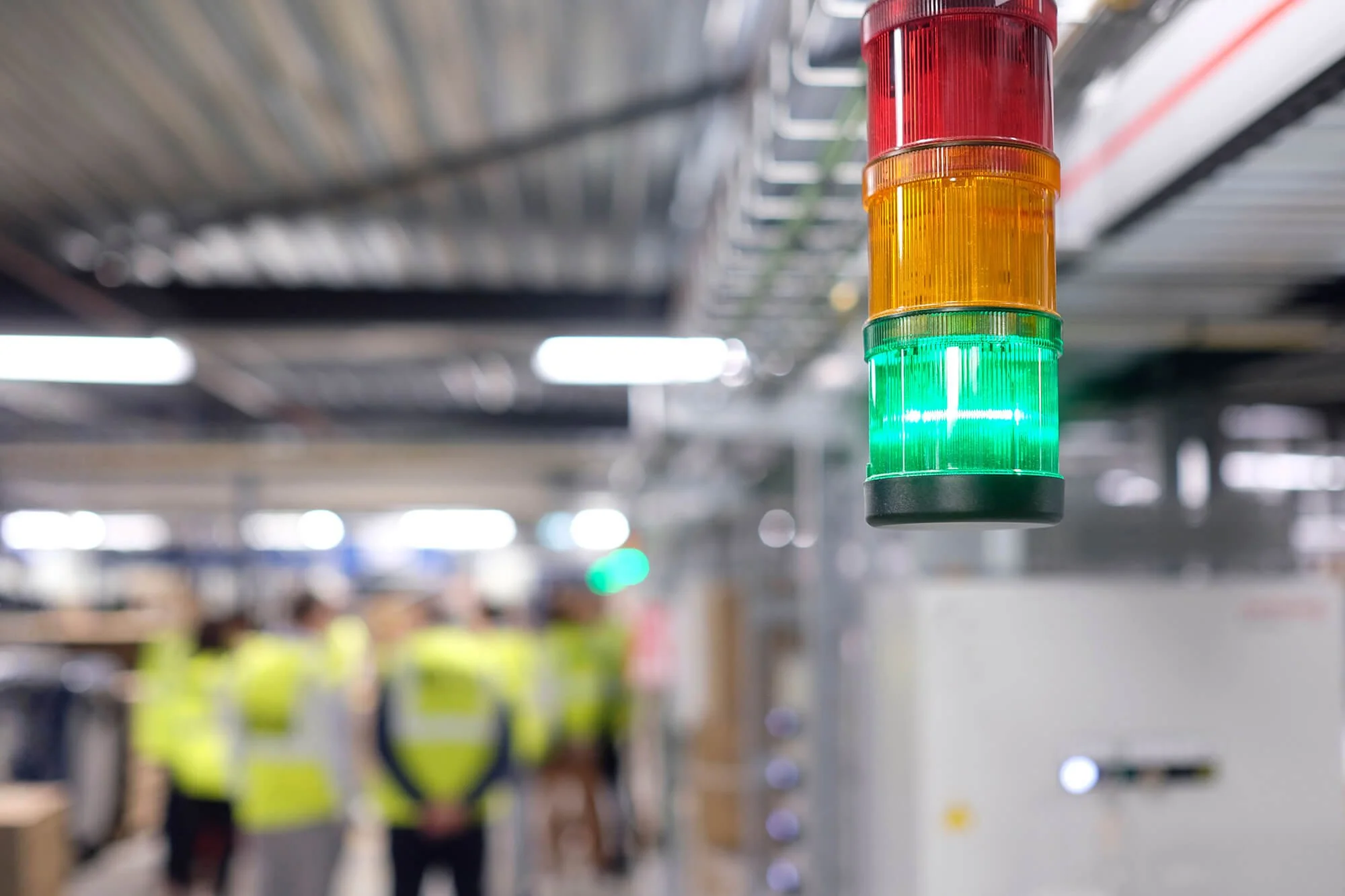 A vertical stack of four LED indicator lights in red, yellow, green, and black, hanging from a ceiling with blurred background of people in yellow safety vests in an industrial or warehouse setting.