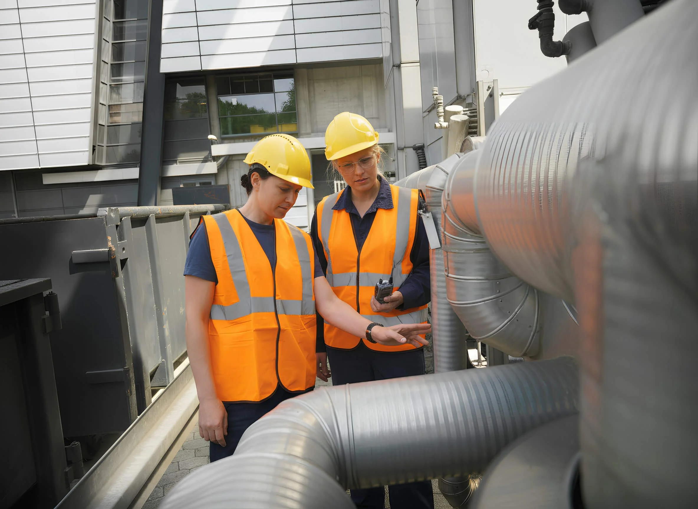 Two women in yellow safety helmets and orange safety vests examining industrial piping outside a modern building.