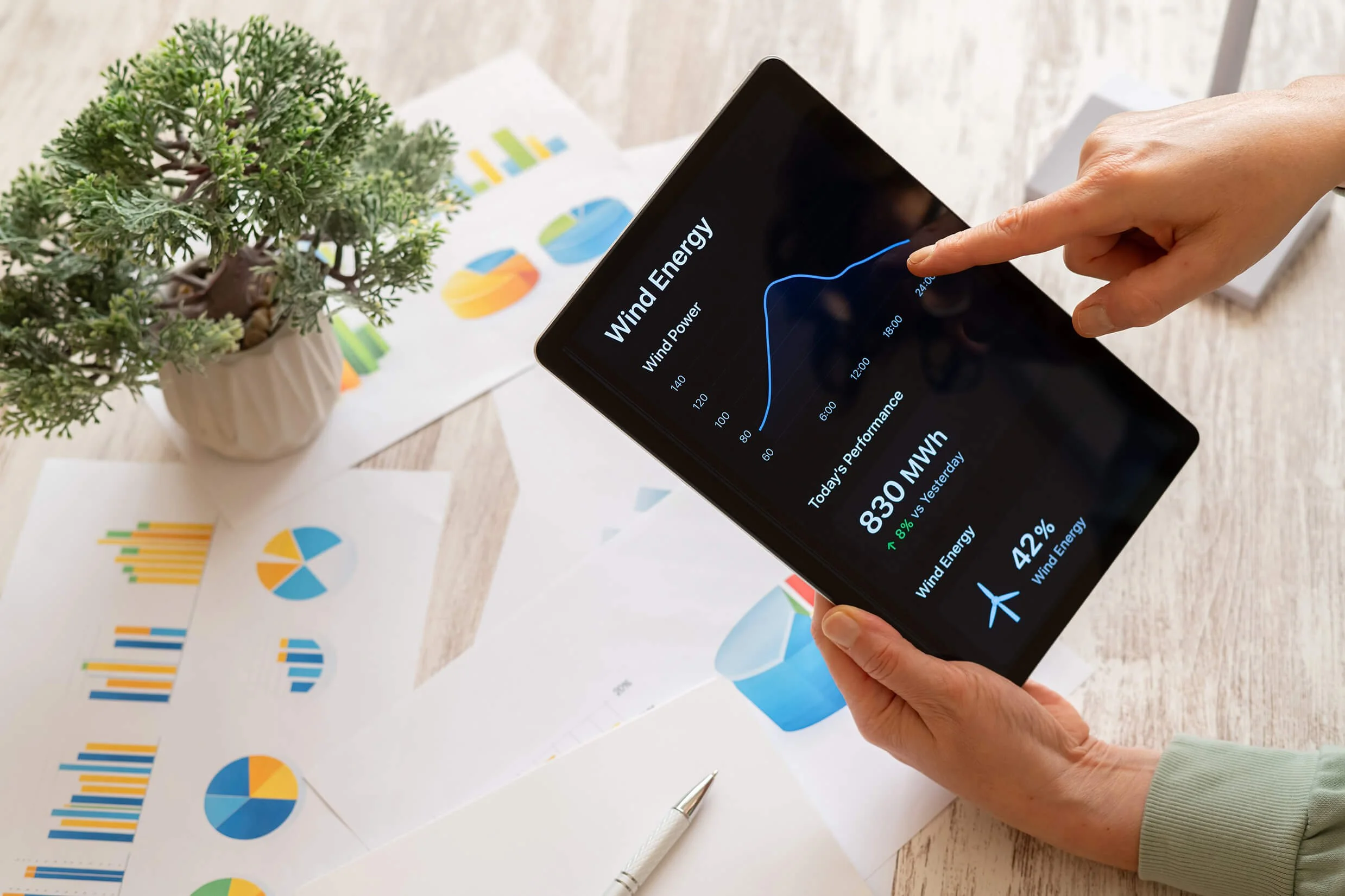 A hand holding a tablet displaying a wind energy chart. The table has printed charts and graphs related to energy data, with a potted plant in the background.