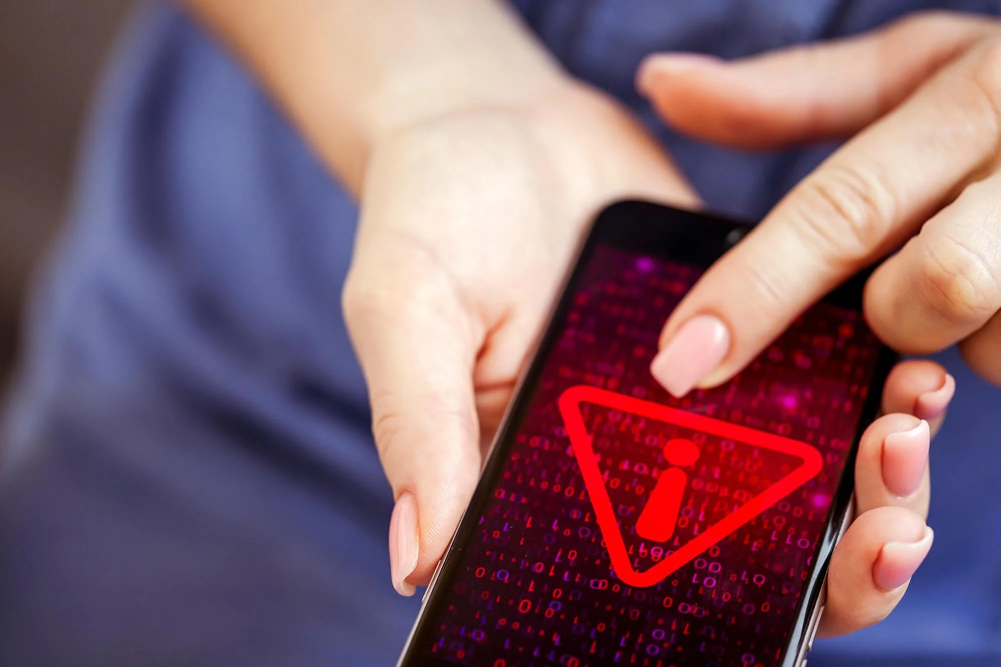 Close-up of a person's hand holding a smartphone displaying a red warning triangle with an exclamation mark and binary code background.