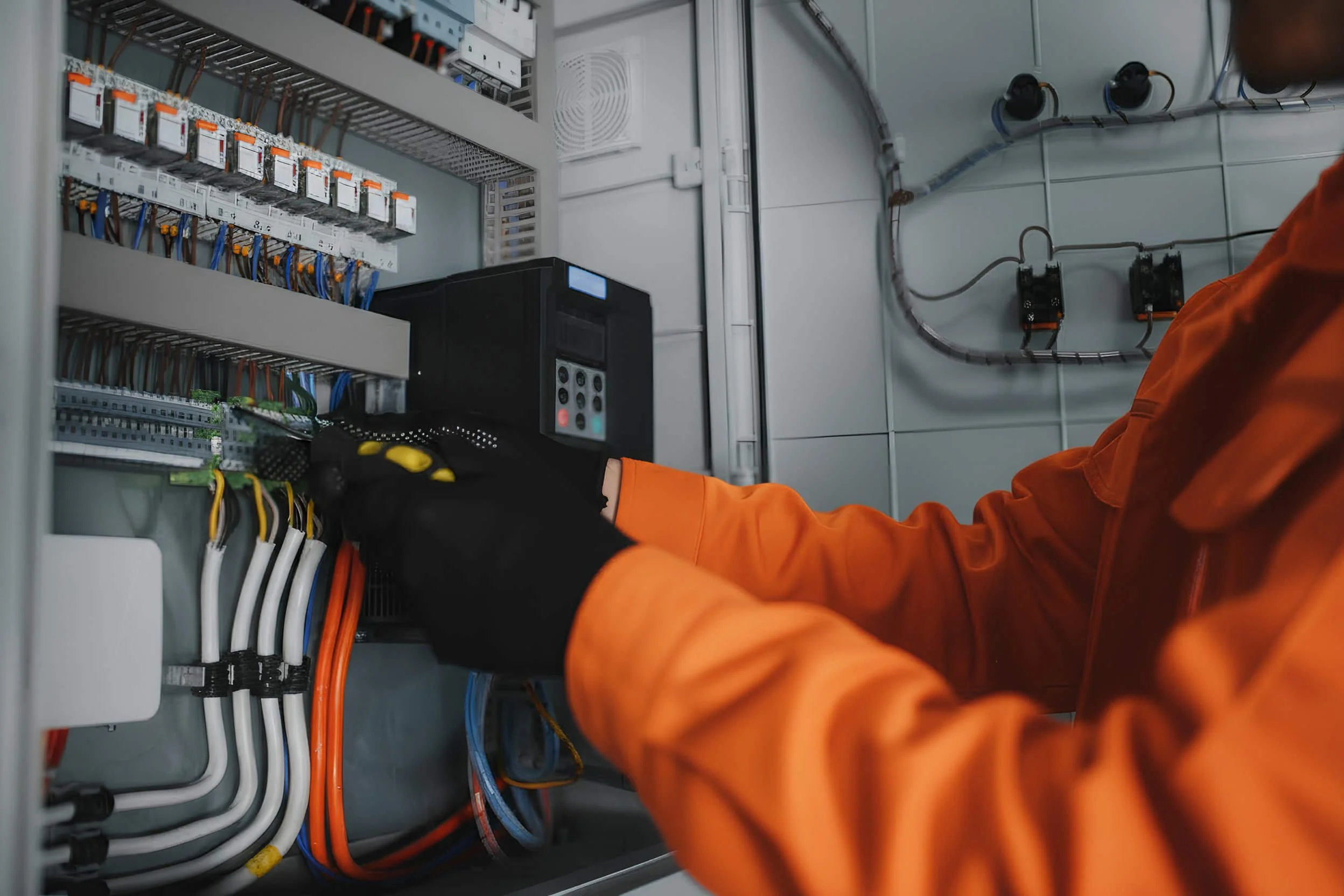 An electrician wearing orange gloves and a matching orange jumpsuit working inside an electrical control panel with various wires and circuit breakers.