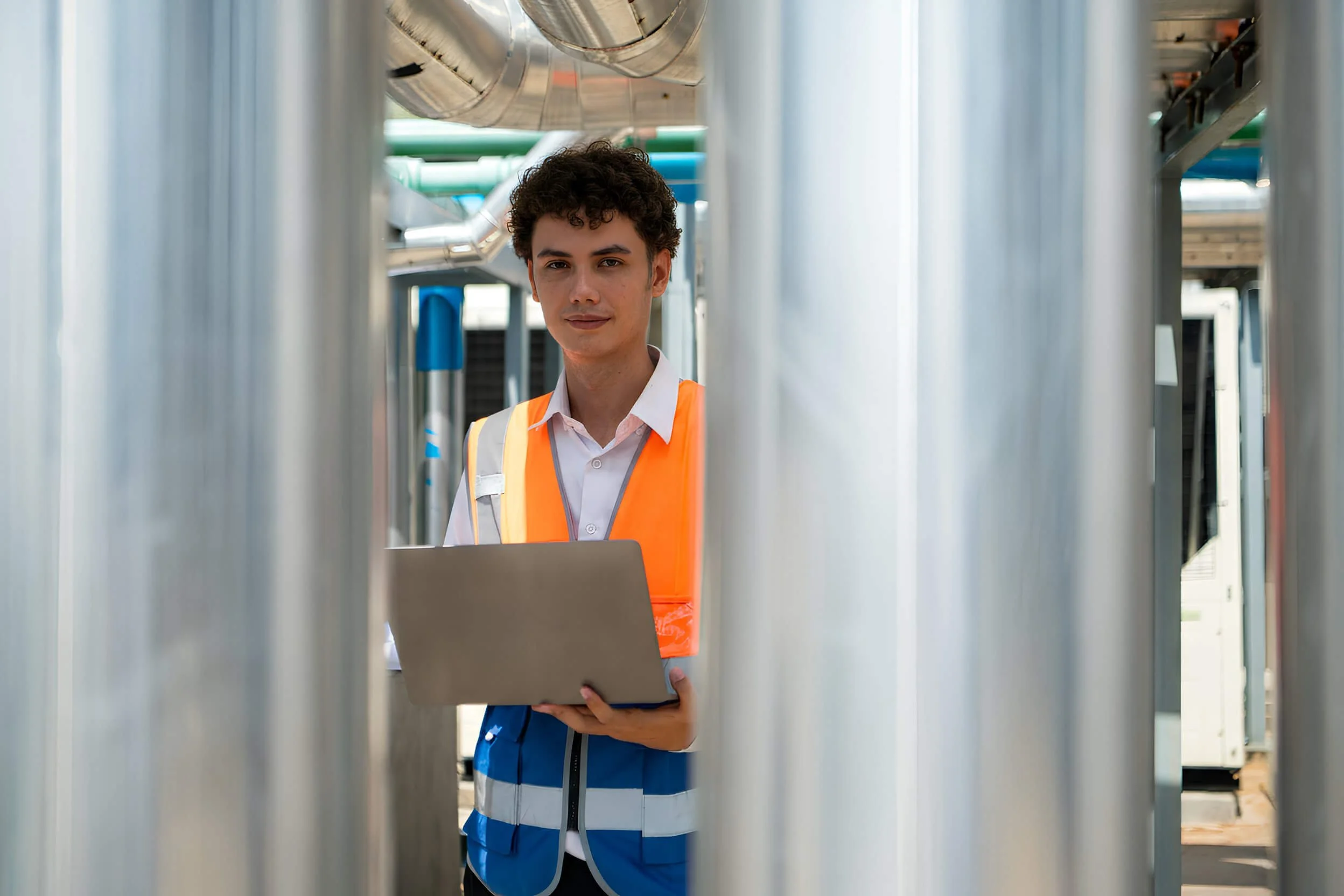 A young man with curly brown hair is working in an industrial or manufacturing setting. He is wearing a white shirt, an orange safety vest, and holding a laptop. The background includes large metallic pipes and machinery.