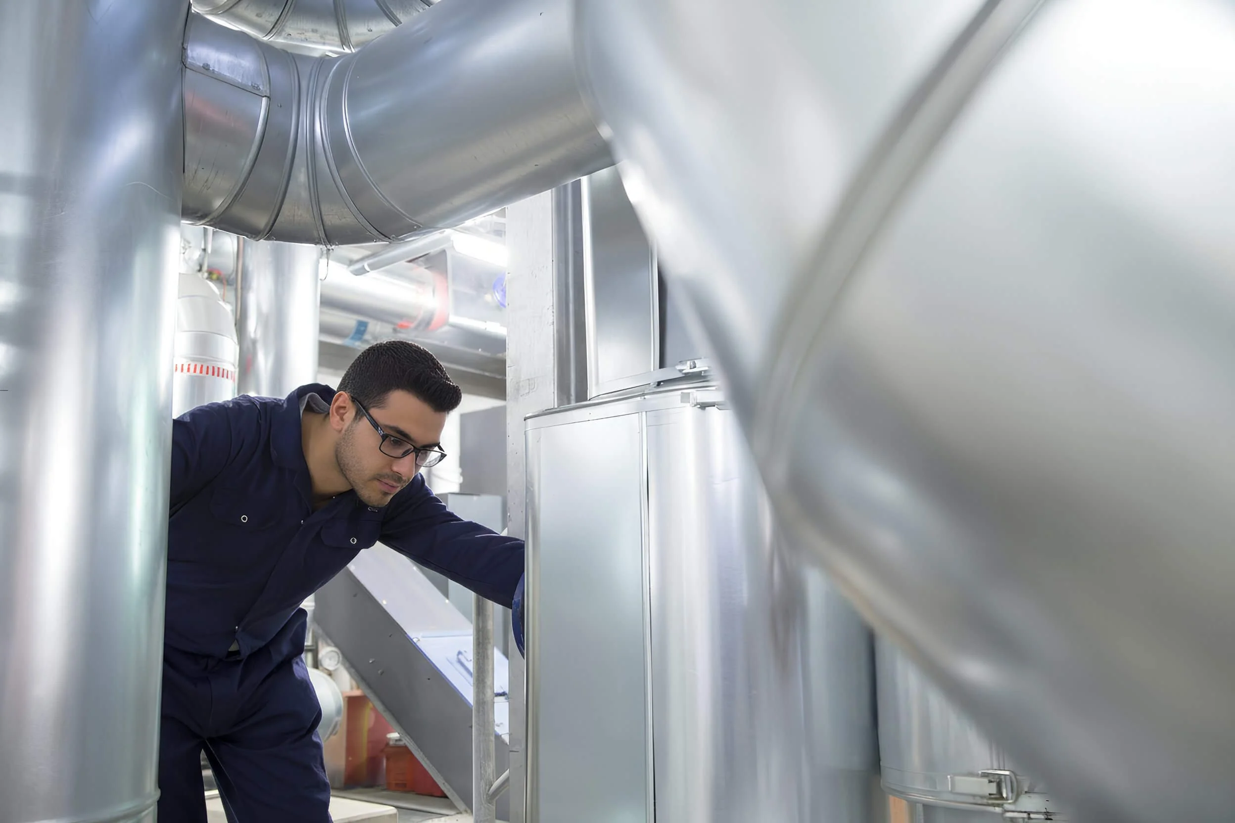 A man wearing glasses and a dark blue uniform working inside a facility with large industrial metallic pipes.