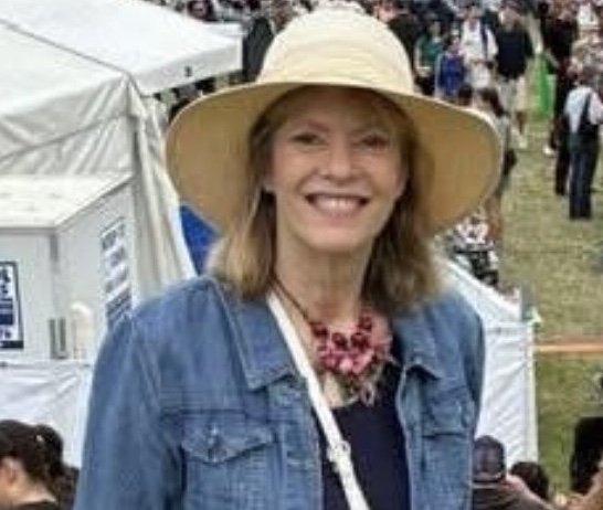 Smiling woman wearing a large sun hat, denim jacket, and pink jewelry at outdoor event with tents and crowd in the background.