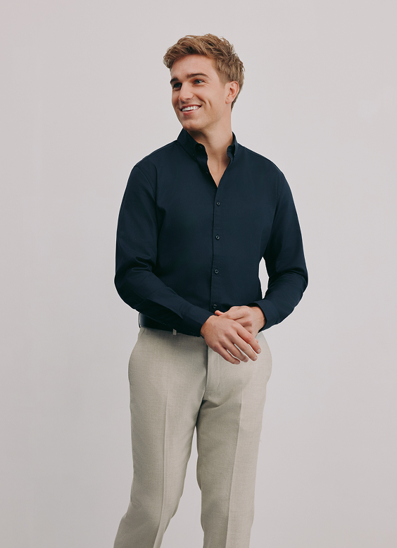 A young man smiling, dressed in a dark blue button-up shirt and light-colored pants, standing against a plain white background.